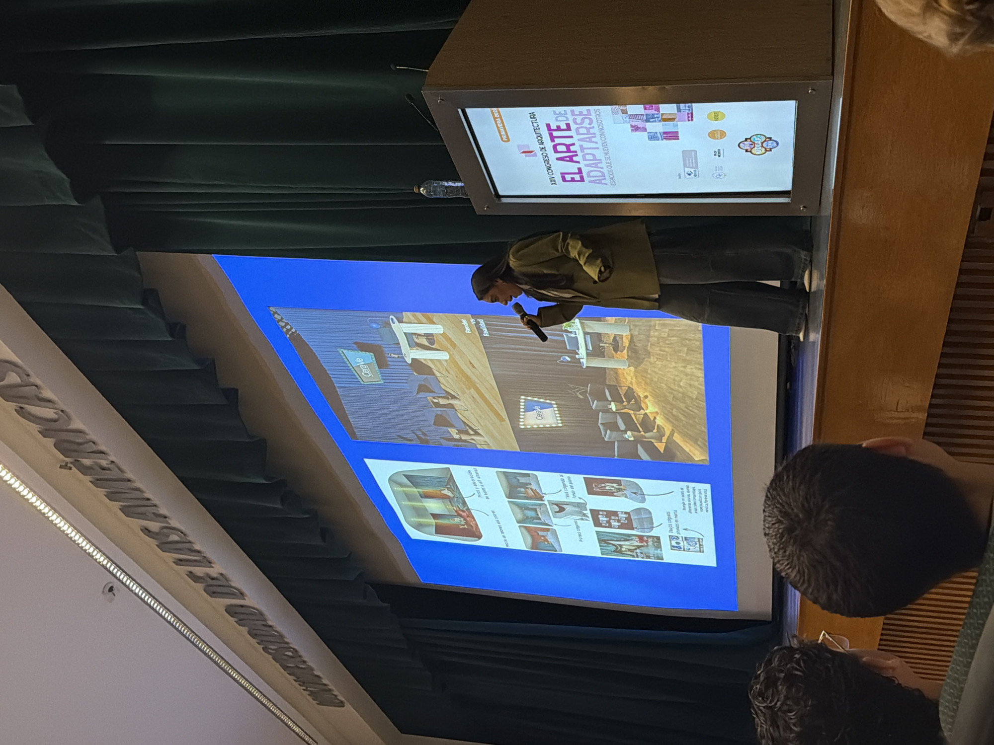 Here is a detailed description of the image:
The image shows a presentation in progress. A person is standing at a podium, speaking into a microphone. A screen behind them displays a presentation titled "EL ARTE DE ADAPTARSE" (The Art of Adapting), part of the "XXIV CONGRESO DE ARQUITECTURA." The presentation includes images of what appears to be interior design concepts, labeled "Props y decoracion," along with text such as "Render VS Realidad" and brands like "Cerave." Some audience members are visible in the foreground. The location appears to be a lecture hall within the "UNIVERSIDAD DE LAS AMERICAS." A water bottle is placed on the podium.