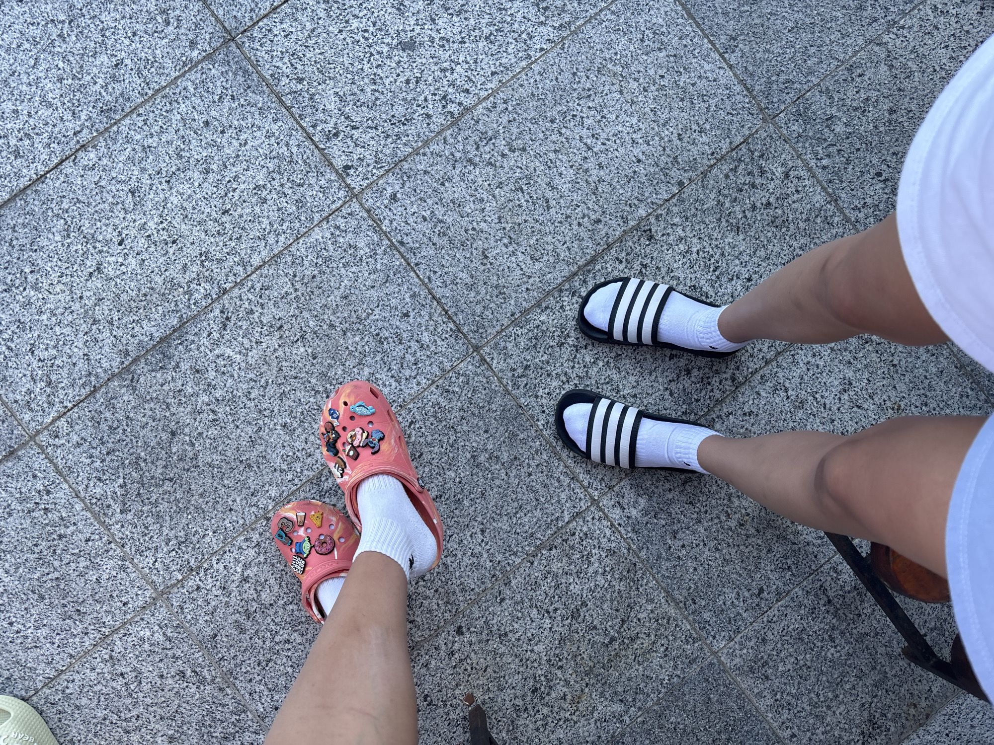 This image shows a low-angle shot of two people's feet standing on a granite tiled surface. One person is wearing pink Crocs adorned with various charms, including cartoon characters, a donut, and a drink. They are also wearing white ankle socks. The other person is wearing black Adidas slides with white stripes, also with white ankle socks. The scene appears to be outdoors, possibly on a sidewalk or patio, given the tiling and the presence of what looks like a metal chair leg in the lower right corner. The lighting suggests it is daytime, with no indication of specific weather conditions. The focus is on the footwear, with the rest of the people and the surroundings out of focus or cropped out. The image captures a casual moment, focusing on fashion and footwear.