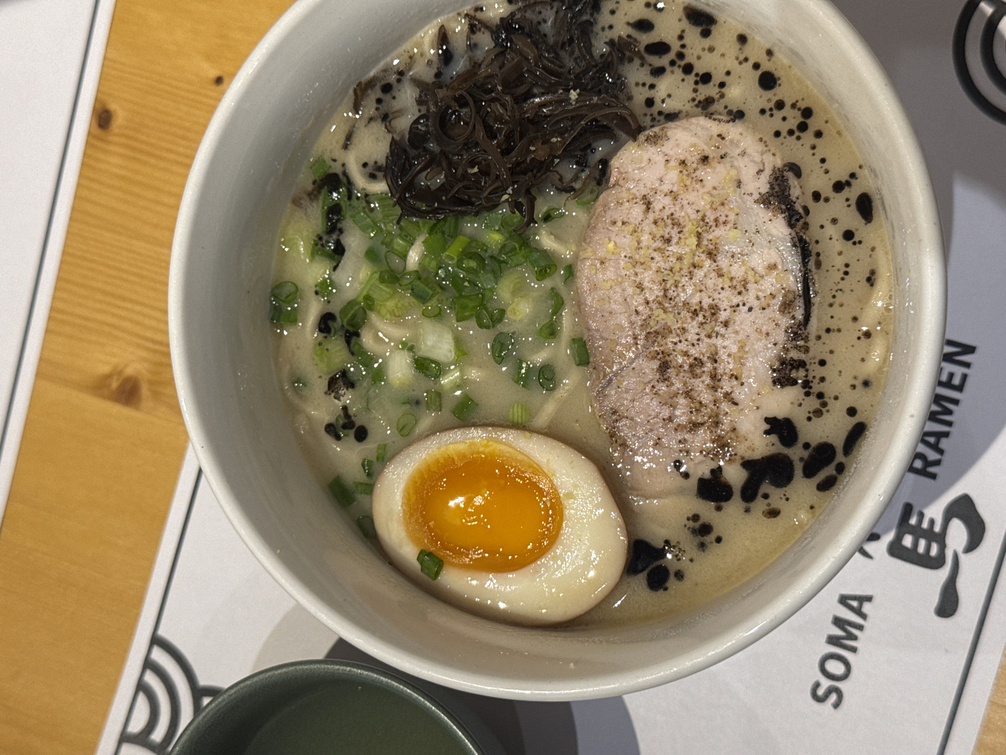 This image shows a bowl of ramen at SOMA Ramen in Tbilisi, Georgia. The ramen contains broth, noodles, a sliced egg with a bright orange yolk, a slice of meat sprinkled with seasoning, green onions, and black fungus. The bowl is white and sits on a placemat with the words "SOMA RAMEN" and stylized Japanese characters. A small green bowl sits beside the ramen bowl. The scene is photographed from directly above.