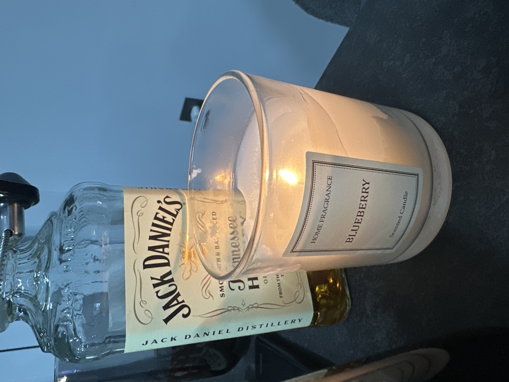 In an indoor setting, a close-up shot focuses on a glass jar candle and a partial view of a Jack Daniel's bottle. The candle, lit and with a visible flame, is in the foreground, its label reading "HOME FRAGRANCE BLUEBERRY Scented Candle." The Jack Daniel's bottle, partially empty, stands behind and to the left of the candle. The bottle's label, a creamy yellow, is clearly visible, featuring the iconic "Jack Daniel's" script logo. The background is a plain, light blue wall. The surface on which the objects rest is a dark, textured material, possibly a countertop or table.