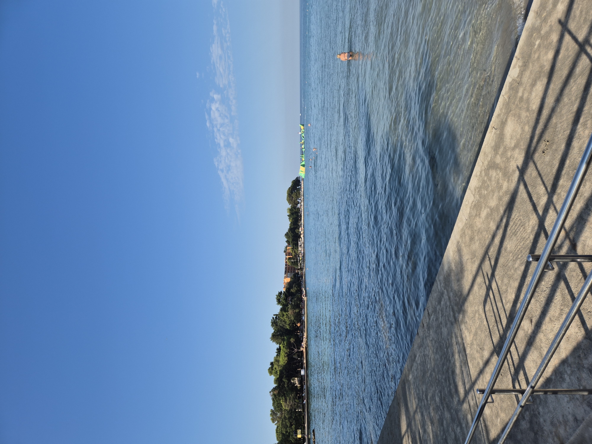 The image shows a view of the Adriatic Sea from a concrete walkway in Monterol, Croatia. The sky is clear and blue with a few wispy clouds on the horizon. The sea is calm and clear, with gentle waves lapping at the shore. In the distance, a line of trees and buildings forms the coastline, with a few people visible on the beach. A few people are in the water, some swimming and one person standing waist-deep in the water. There is an inflatable water park in the middle distance. The concrete walkway in the foreground has metal railings and is casting shadows from an unseen light source. The overall scene is one of a peaceful summer day at the beach.