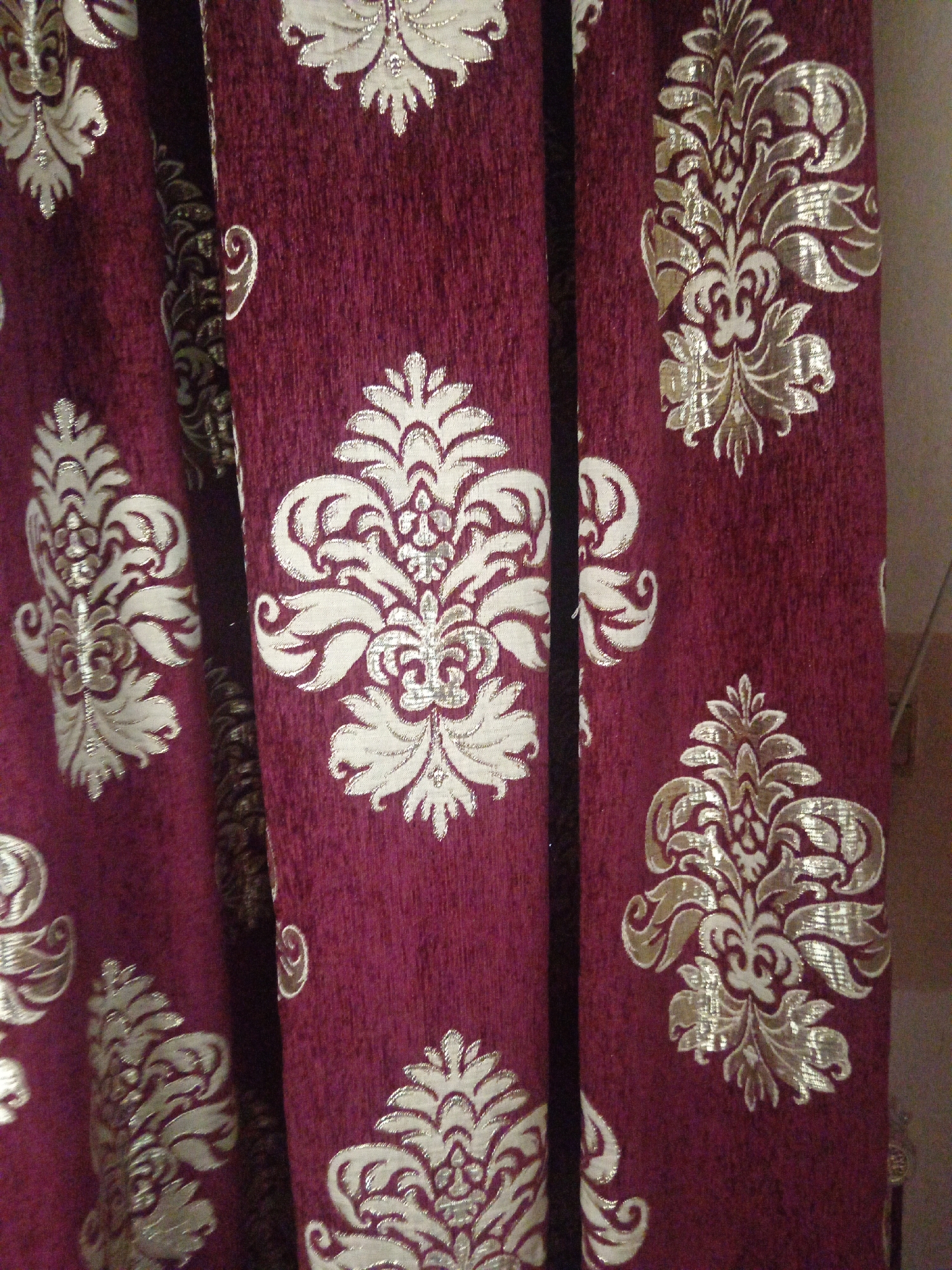 The image shows a close-up view of burgundy-colored curtains with a damask pattern. The pattern is a repeating motif of ornate floral designs in a metallic gold or silver color. The curtains are gathered or folded, creating vertical lines and shadows that add depth to the image. The texture of the fabric appears to be plush or velvet-like, with a slight sheen from the metallic threads. The background is out of focus but appears to be a wall with a light color. There are no people or animals visible in the image. The scene is indoors, likely a room with windows covered by these curtains. The location is not specified beyond "Unknown, Unknown."