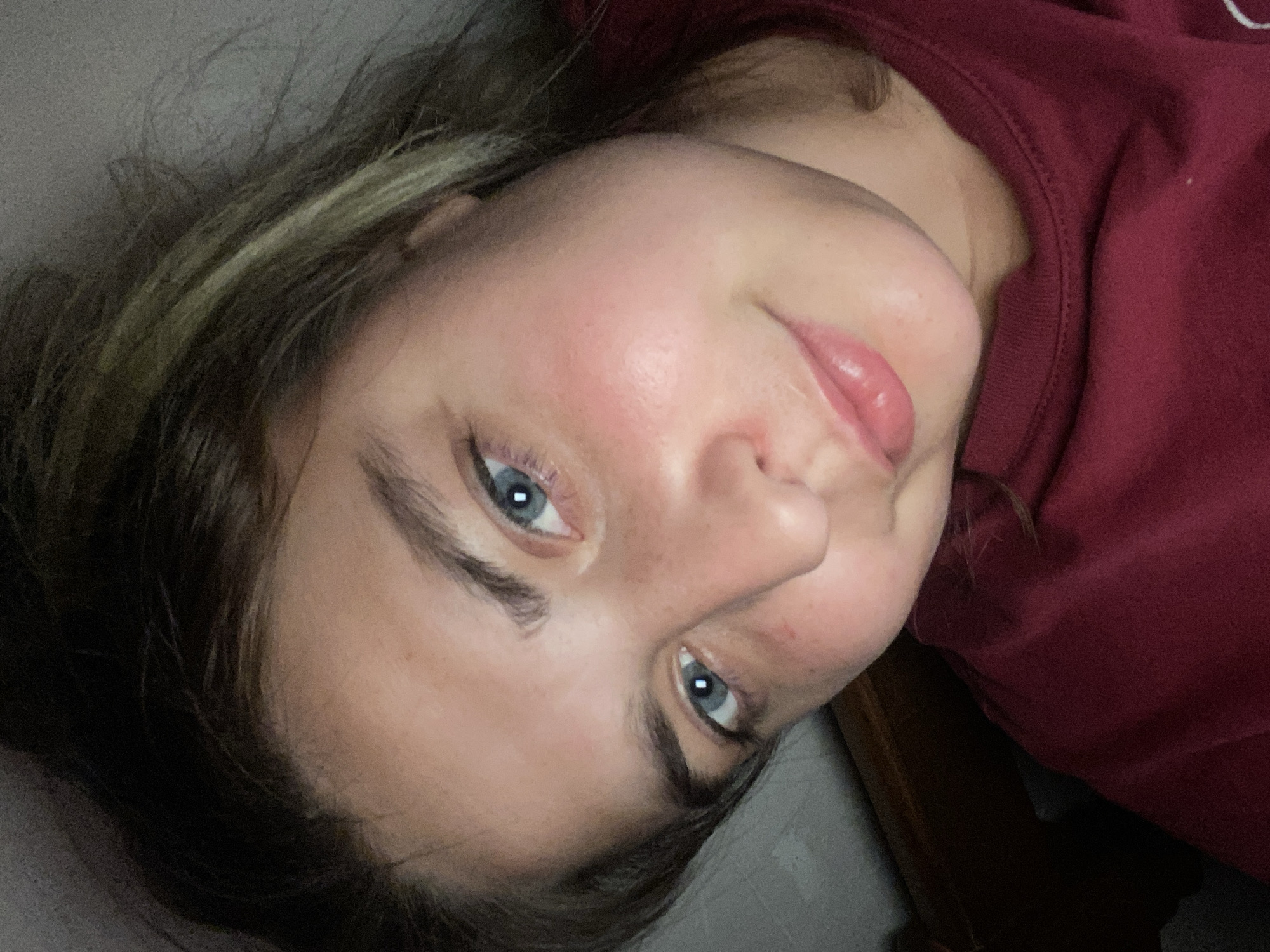 The image is a close-up, low-angle selfie of a young woman with light skin, rosy cheeks, and freckles. She has blue eyes and brown hair with blonde highlights, which is pulled back. She is wearing a dark red t-shirt. The background is dimly lit and appears to be indoors, possibly in a bedroom, with a hint of a wooden headboard visible. There is no indication of the specific location within the city of Otradnaya, Russia. The lighting is soft, and the focus is on her face.