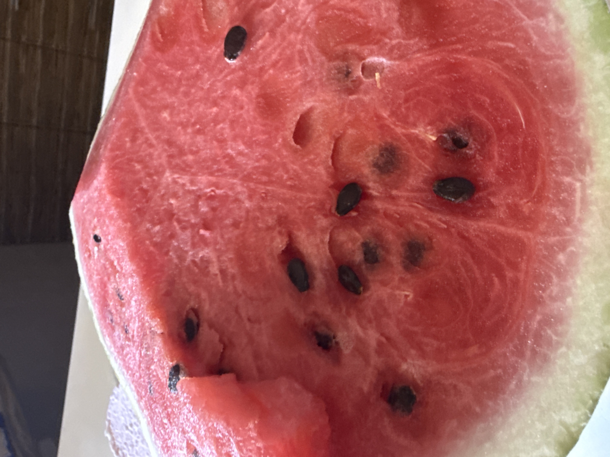 A close-up, top-down shot shows a slice of ripe watermelon. The vibrant red flesh is dotted with numerous black seeds, some of which are partially submerged within the fruit. A bite has been taken out of the upper left portion of the slice, revealing a softer, pinker interior. The rind, a pale green and white, is visible along the bottom edge of the image. The background consists of a textured, brown surface and a white surface, suggesting an indoor setting.