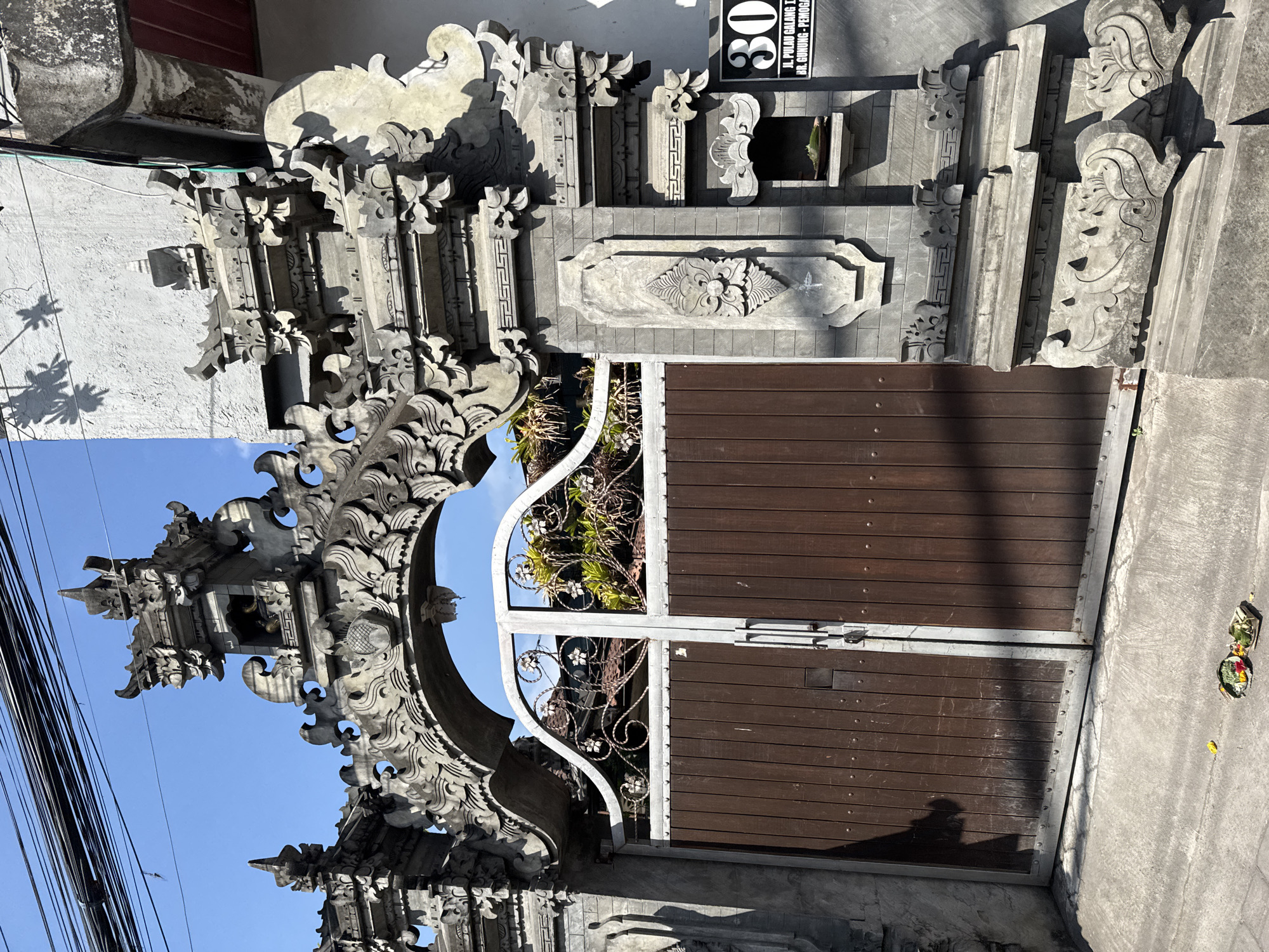 The image shows a traditional Balinese stone gate and wooden doors located at an address in Denpasar, Indonesia. The gate is elaborately carved with intricate details, typical of Balinese architecture, and features a large arched opening. The wooden double doors are dark brown and appear to be made of vertical planks. Above the doors, a metal grille with decorative floral elements is visible.
On the right side of the gate, a house number sign with "30" is prominently displayed, along with a street address reading "JL. PULAU GALANG I, BR. GUNUNG - PEMOGAN". Below the house number, there is a small niche in the stone structure containing a leaf and what appears to be a small offering.
The ground in front of the gate is paved with concrete. A small, colorful offering, likely a *canang sari*, is placed on the ground near the left door. In the background, lush green foliage is visible through the top portion of the gate. Overhead, a tangle of electrical wires is strung against a clear blue sky. The overall scene depicts a residential entrance with cultural elements and functional components.