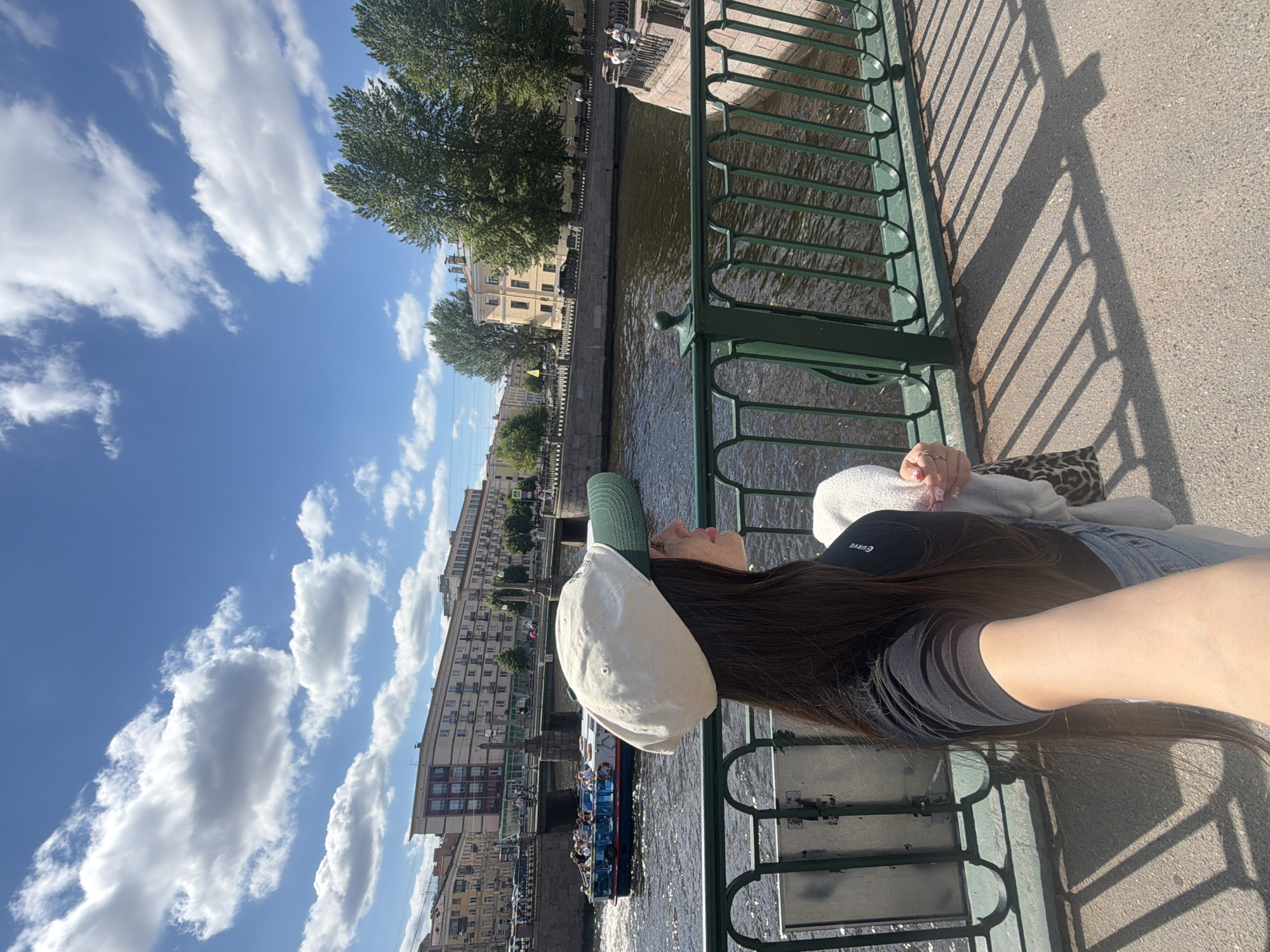 A woman with long dark hair, wearing a black t-shirt, jeans, and a light-colored baseball cap with a green brim, stands on a bridge in Saint Petersburg, Russia, taking a selfie. She is holding a white towel and a bag with a leopard print strap. In the background, a boat with passengers cruises on a canal, lined with trees and historic buildings. The sky is blue with fluffy white clouds. The bridge has a green metal railing, and shadows of the railing are cast on the paved walkway.