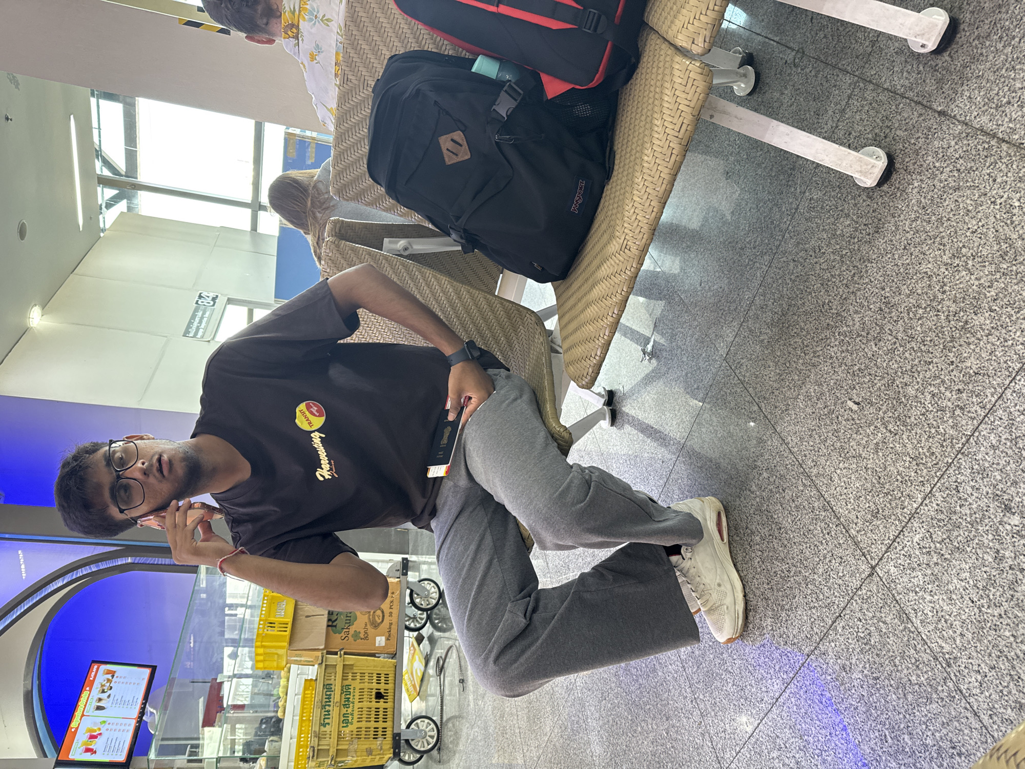 In Mai Khao, Thailand, a young man with dark hair and glasses sits on a woven bench in what appears to be an airport or transit lounge. He is wearing a dark t-shirt with a graphic print and grey sweatpants, and his right hand is raised to his ear as he talks on a rose-gold colored phone. A boarding pass is held in his left hand. Beside him on the bench is a black Jansport backpack. Another backpack, red and black, is partially visible to the right. The background shows an illuminated menu board displaying drinks and food items, stacks of yellow plastic crates, and a large window with daylight streaming in. The floor is a speckled grey tile. The man's white sneakers are visible.
