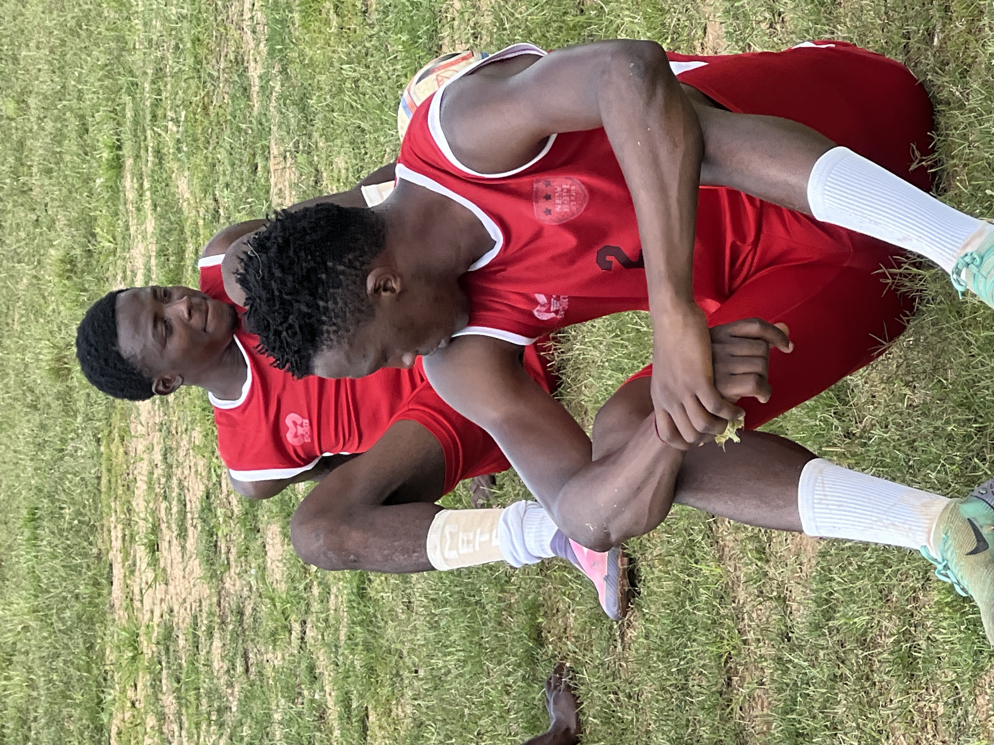Two young Black men in red athletic uniforms are sitting on a grassy field, appearing to be on a break during a game or practice. The man in the foreground is facing to his left, looking down and holding a small yellow object in his right hand. He is wearing a red sleeveless jersey with white trim and a crest featuring three stars and text, and red shorts. His legs are crossed, and he has a white athletic sock pulled up to his calf, paired with a green and black Nike cleat. A white bandage or compression sleeve is visible above the sock on his left leg. The man in the background is facing to his right, looking forward with a slight smile. He is also wearing a similar red athletic uniform. The ground is covered in short, dry grass with patches of exposed dirt. The environment suggests an outdoor sports facility, likely in Jos, Nigeria, given the context. The overall lighting indicates it is daytime, likely under natural sunlight.