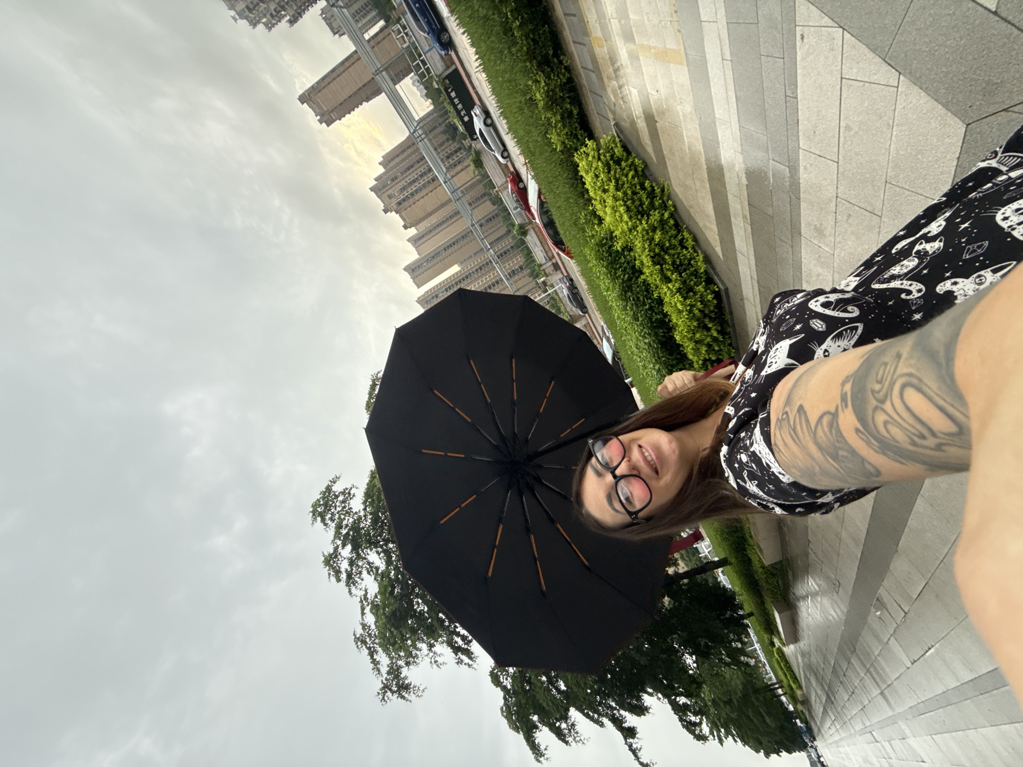 A woman is taking a selfie outdoors in Huizhou, China, holding an open black umbrella above her head. She is wearing glasses and a black and white patterned dress. Her left arm, which is heavily tattooed, is extended towards the camera. The background shows a tree-lined street with a row of cars, green bushes, and tall apartment buildings under a cloudy sky. The pavement appears wet, suggesting it may have recently rained.