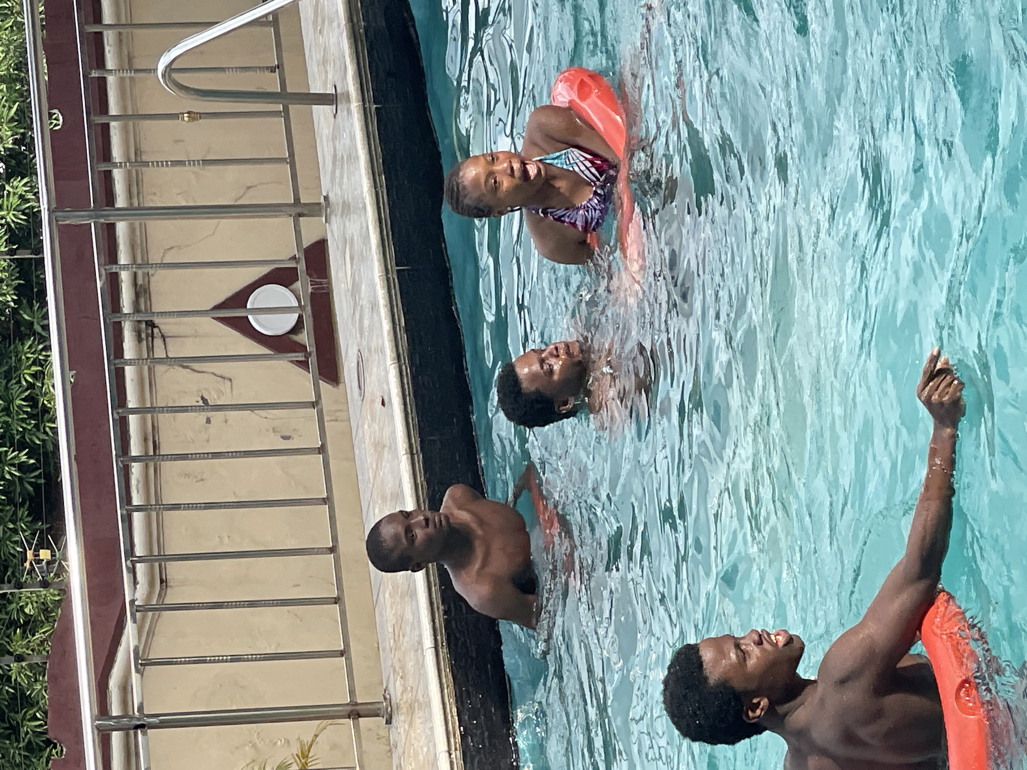 In Nkpor, Nigeria, four young Black individuals are enjoying a sunny day at a swimming pool. Three males and one female are in the crystal-clear blue water. The female, positioned at the top of the frame, wears a colorful bikini and a bright orange inflatable ring around her waist. She smiles at the camera, her face illuminated by the bright sunlight. Just below her, a male is partially submerged in the water, his face turned upwards. Further down, another male, with dark skin and short hair, is in the water, with a reddish-orange ring visible. He is reaching upwards with his right arm, his fingers pointed towards the sky, as if playing or interacting with someone outside the frame. The fourth individual, a young man with dark skin and short, curly hair, is seen at the bottom right of the frame. He has a bright orange inflatable ring around his waist and is looking to his left. The pool area has a tiled edge, and a metal railing with a handrail is visible along the side. Behind the railing, there is a light-colored wall with some decorative elements. Green foliage from trees is visible in the background to the left, suggesting a warm and pleasant day. The overall mood appears joyful and carefree, with the subjects actively engaging in pool activities.