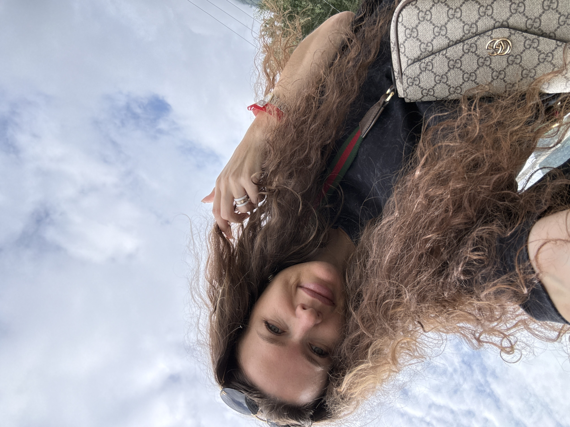 The image, taken from a low-angle perspective, shows a woman with long, curly brown hair, wearing a black t-shirt and sunglasses perched on her head. She is also wearing rings on her hand and a red bracelet. A handbag with a distinctive monogram pattern and a red and green stripe is visible across her chest. The background consists of a cloudy sky and some foliage. The overall impression is of a candid outdoor photograph, possibly a selfie. The metadata for this image states it was taken in Chemal, Russia.