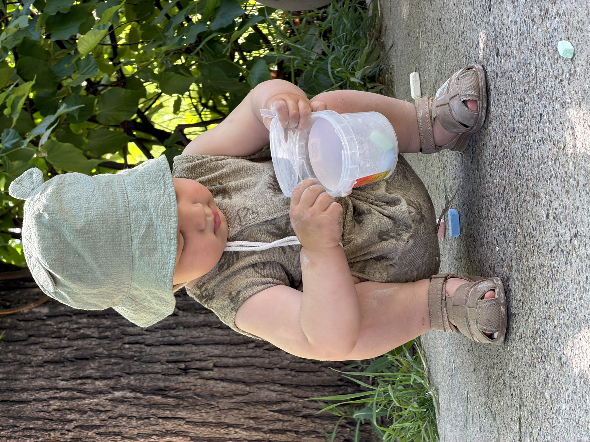 A baby wearing a light green hat and a matching brown onesie sits on a concrete surface, possibly a sidewalk, outdoors. The baby is holding a clear plastic container with a white lid, seemingly examining its contents. Several pieces of colored chalk are scattered around the baby on the concrete. The background includes a tree trunk, green vegetation, and some foliage. There is no indication that this image was taken in Chaikovsky, Russia.