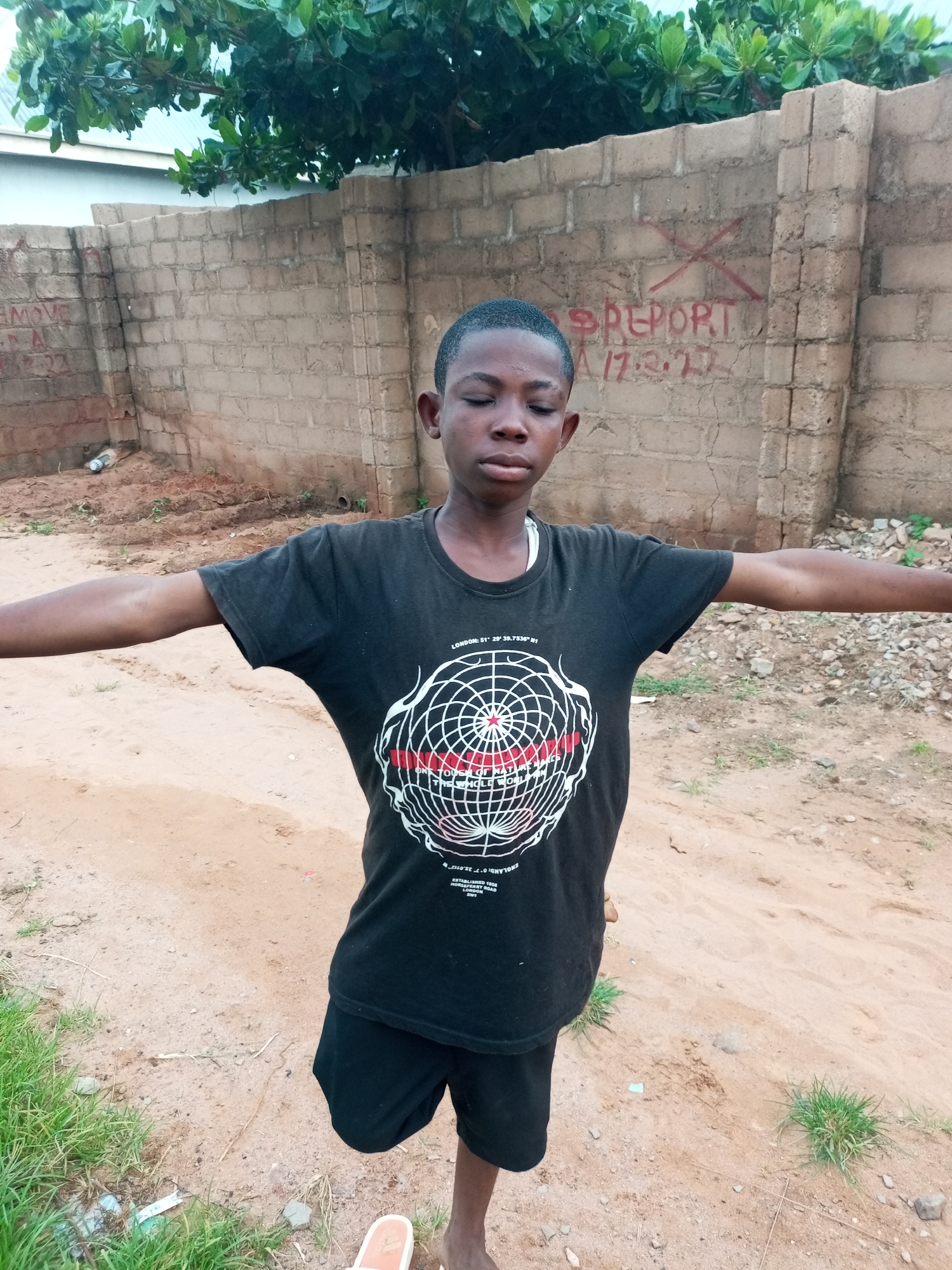 A young Black male is standing outside with his eyes closed and arms outstretched in front of a brick wall. He is wearing a black t-shirt with a graphic design on it and black shorts. The wall behind him has graffiti on it, including the words "REPORT" and a date. The ground is dirt and there is some grass and vegetation around. The image appears to be taken in a developing area, possibly in a rural or semi-urban location in Tempure, Nigeria.