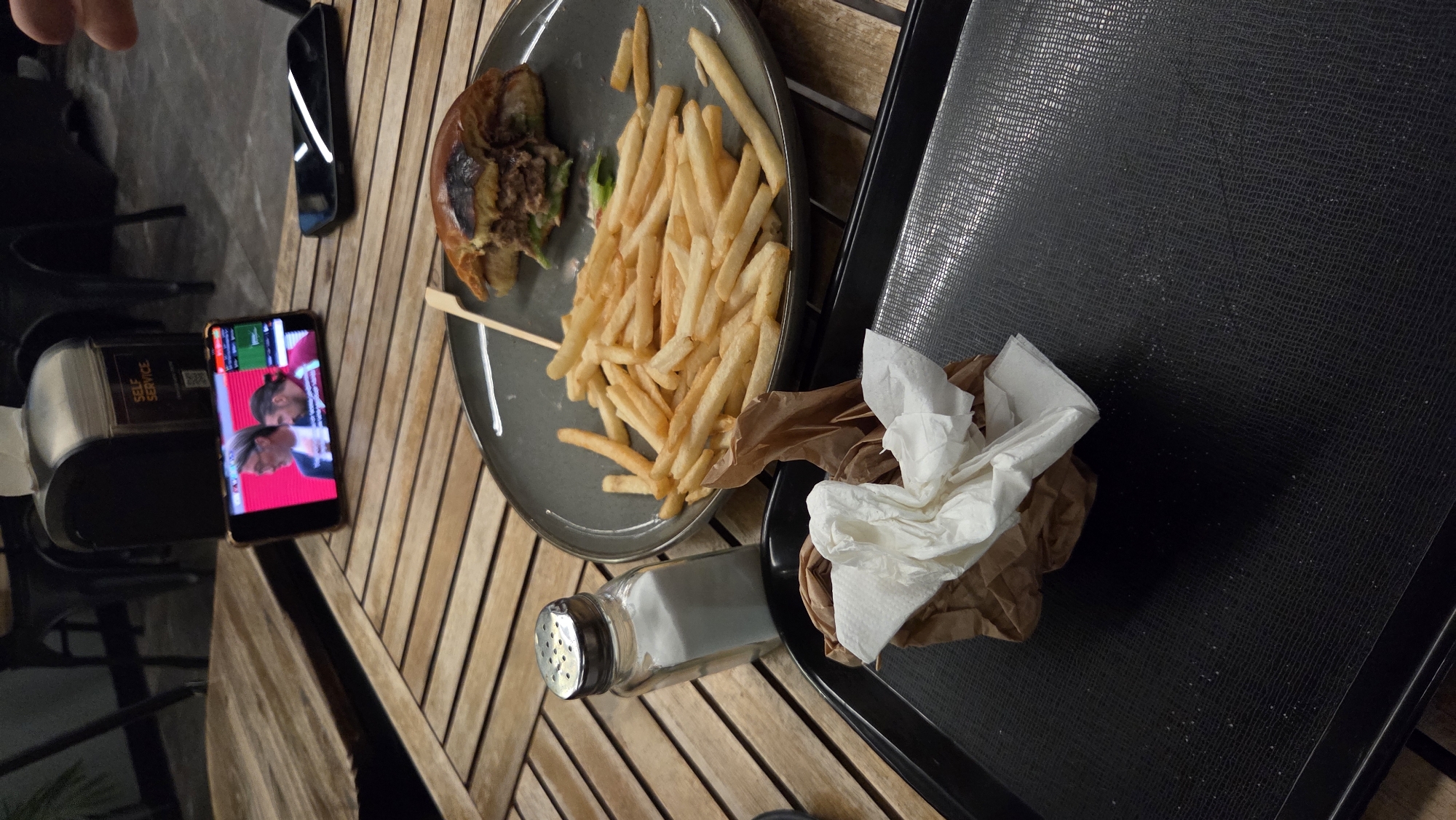 A partially eaten burger and a pile of french fries sit on a dark plate on a wooden table. A salt shaker is to the left of the plate, and a crumpled paper napkin is on a black tray in the foreground. In the background, a smartphone displays a soccer match. The scene appears to be set in a restaurant in Bansko, Bulgaria.