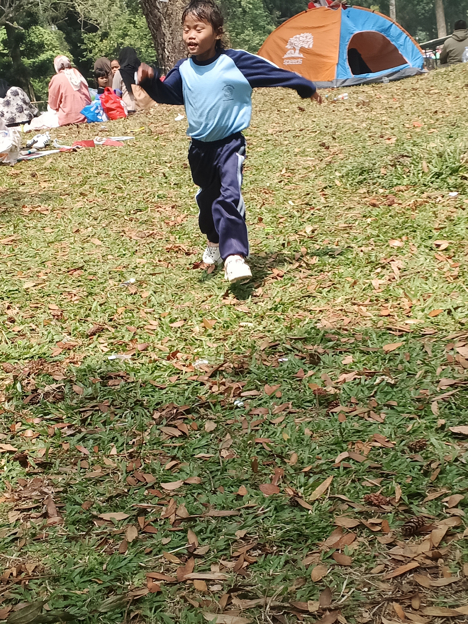 The image shows a child running on a grassy field with fallen leaves. In the background, there are several people sitting on the grass, a tent, and trees. The scene appears to be a park or recreational area. The child is wearing a blue shirt and dark pants, and is in motion, with arms outstretched. Some of the people in the background are wearing headscarves. The tent is orange and blue. The environment is outdoors, with sunlight filtering through the trees, casting shadows on the grass. Based on the provided context, this scene is located in Perbawati, Indonesia.