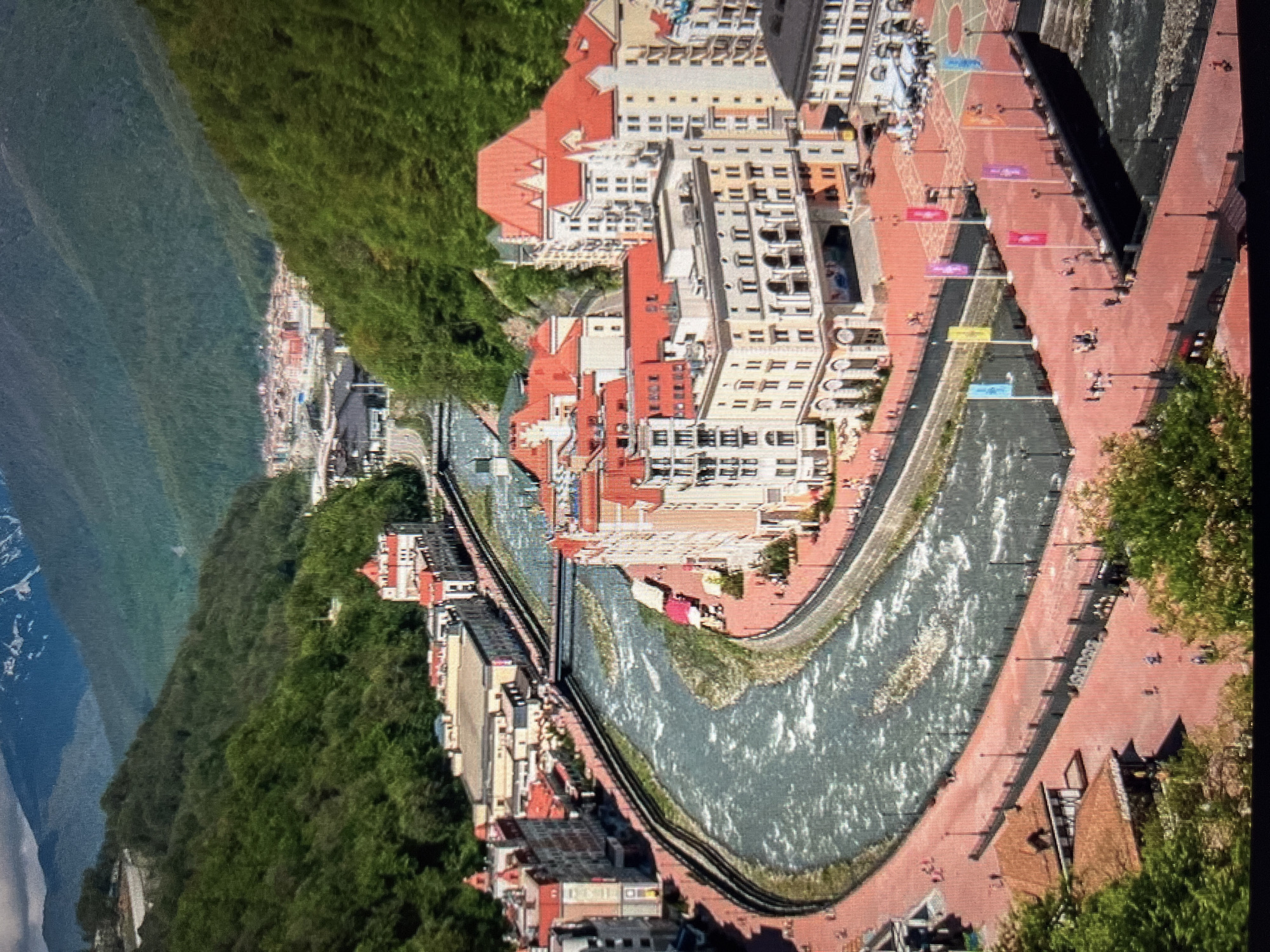 The image shows a scenic view of a resort town nestled in a valley surrounded by lush green mountains, with snow-capped peaks visible in the distance. A wide, fast-flowing river cuts through the town, with a picturesque promenade running along its banks. The architecture features buildings with red tiled roofs and light-colored facades, indicative of European alpine style. There are people strolling along the promenade, some seated at outdoor cafes, and flags lining a pedestrian bridge over the river. The scene suggests a vibrant tourist destination, potentially a ski resort in warmer months. The location is identified as the city of Khimki, Russia, though the visual characteristics strongly resemble a mountain resort rather than a typical urban environment in Khimki, which is located near Moscow.