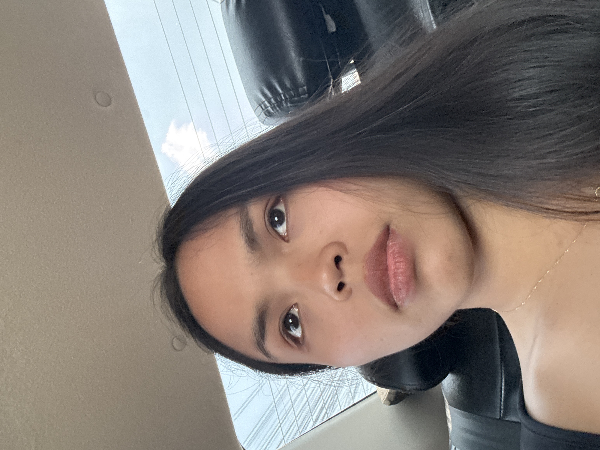 The image shows a young woman with long, dark hair, brown eyes, and fair skin. She is sitting in a vehicle, presumably a car, with black leather seats. The background shows a window with visible power lines and a cloudy sky, suggesting an urban environment. Given the user's prompt, the location is likely Davao City, Philippines. She is wearing a black top and a delicate gold necklace. The angle of the photo is from below, looking up at her face.