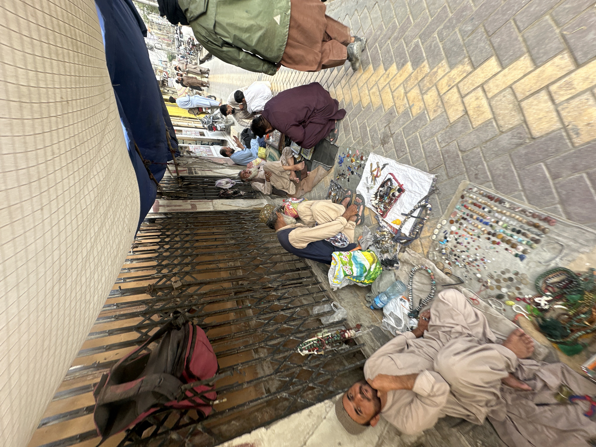 The image shows a street scene in Quetta City Tehsil, Pakistan. Several men are seated or lying on the sidewalk, with items for sale spread out in front of them. These items appear to include beads, jewelry, and other trinkets. The men are dressed in traditional Pakistani clothing, and the background features buildings and other urban elements. The scene is viewed from a slightly elevated perspective, and the angle is tilted.