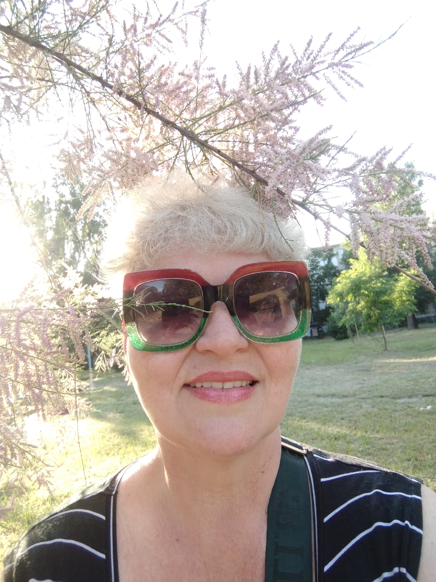 A direct, eye-level selfie captures an adult female situated outdoors in Horishni Plavni, Ukraine. The subject, with short, light-colored hair, wears oversized square-framed sunglasses featuring distinct red and sparkly green sections, with a thin branch from a flowering plant resting across the left lens. She presents a faint smile, her lips lightly parted, and is dressed in a black sleeveless top with thin white horizontal stripes. Directly above and framing her head are numerous branches covered in small, light pink/purple blossoms, characteristic of a Tamarix tree or shrub. The scene is illuminated by strong natural sunlight originating from behind and to the left of the subject, causing lens flare and backlighting. The immediate background features a sun-drenched grassy area with scattered trees, and in the mid-distance on the right, a multi-story building is visible through the foliage.