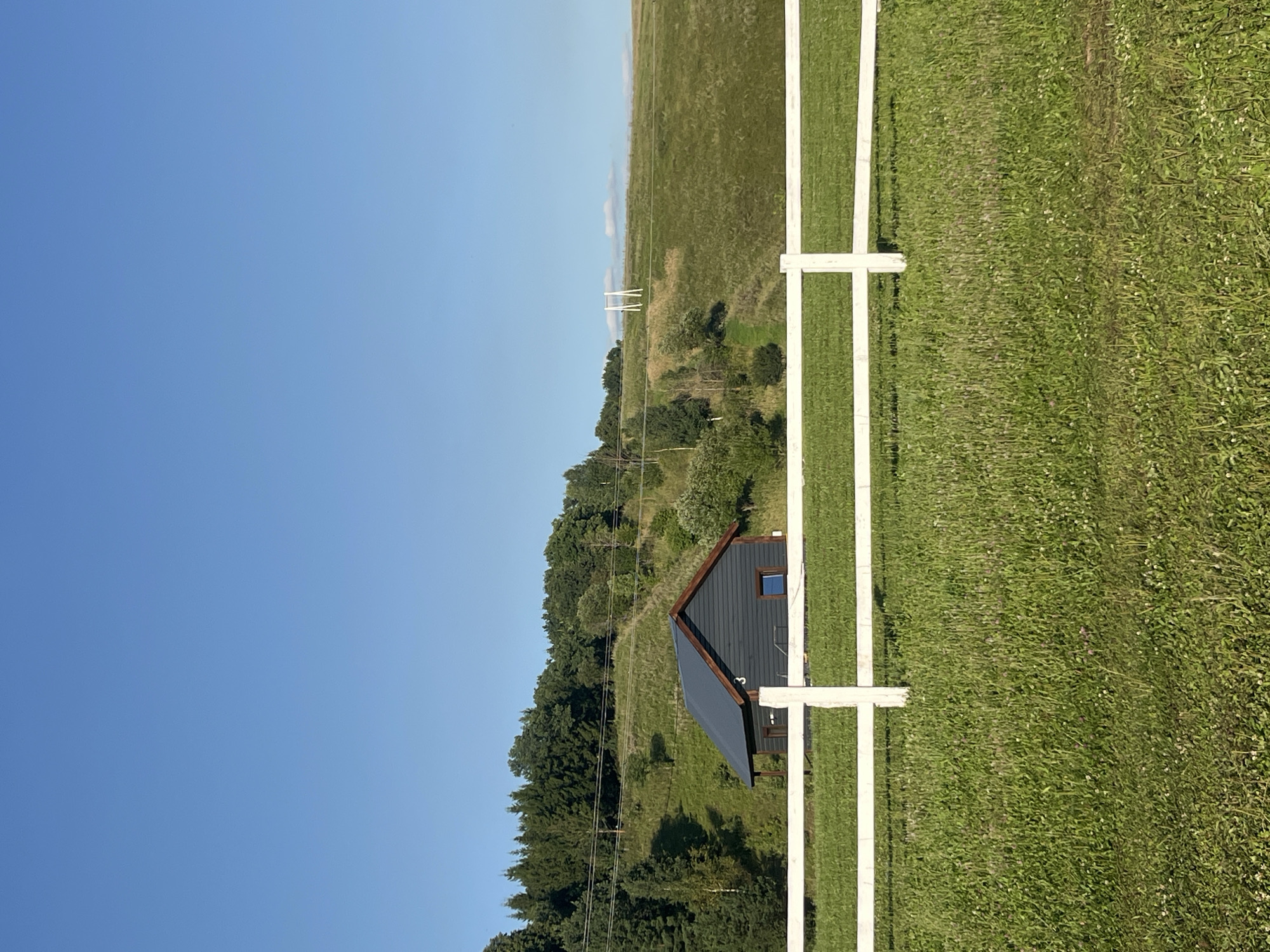 The image depicts a tranquil rural landscape under a clear blue sky. In the foreground, a white wooden fence runs horizontally across the frame, separating a vibrant green grassy field from the scene beyond. Behind the fence, a small, dark gray house with a dark roof and wooden trim is nestled on a gently sloping green hillside. The house has a single window visible on its side and the number "3" is painted on its wall. The hillside is dotted with trees and patches of greenery. In the distance, a line of evergreen trees forms a dense forest along the crest of the hill. Further back, two white, tall, A-frame structures, possibly part of a decorative archway or a landmark, stand against the horizon. The overall impression is one of peace and natural beauty in a rural setting, possibly in or near Srednie Pinyachi, Russia.