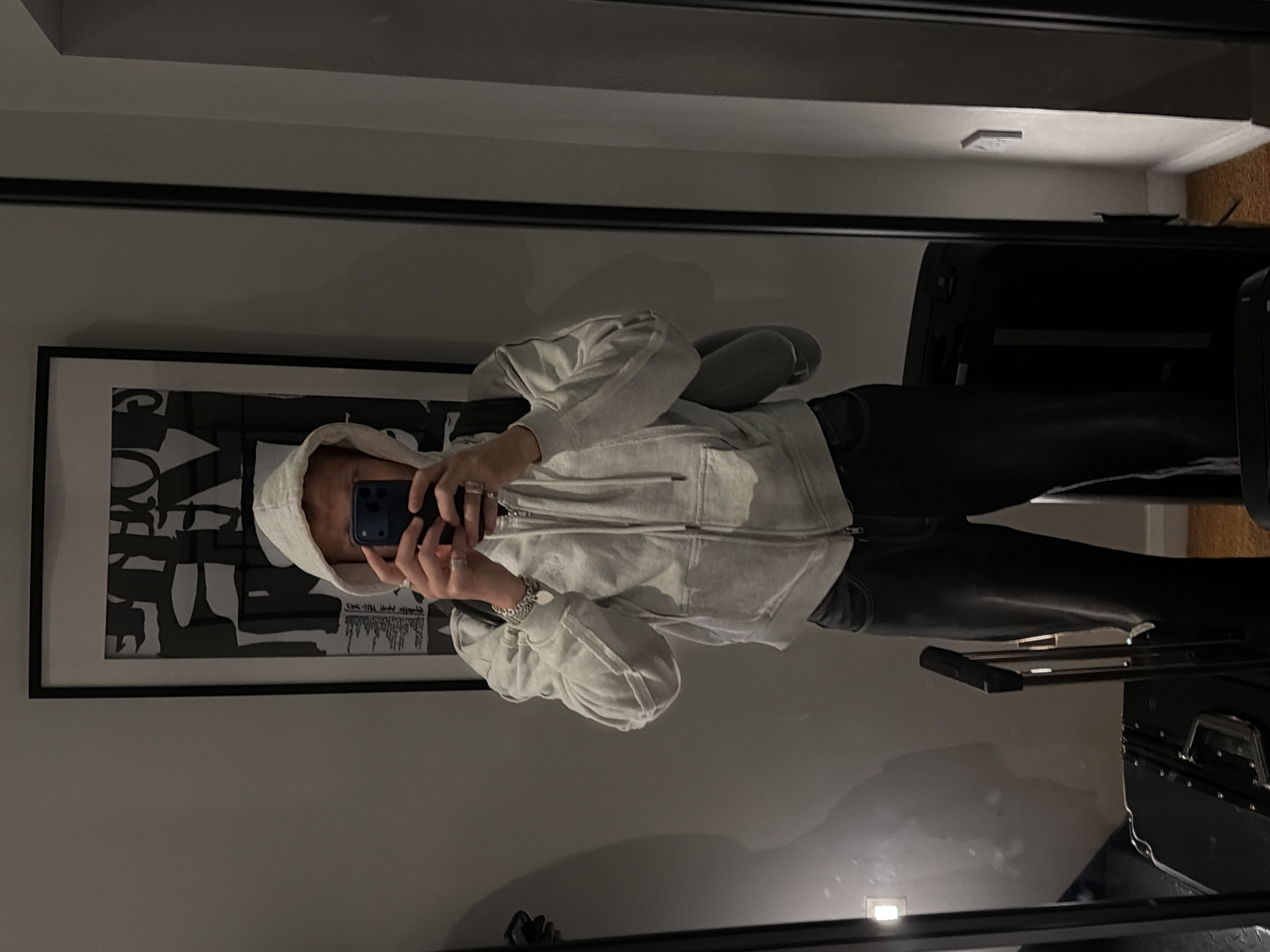 A person, wearing a light-colored hooded sweatshirt, dark trousers, and jewelry on their hands and wrist, stands in front of a mirror, taking a selfie with a smartphone. The person has a backpack on. In the background, a framed abstract artwork hangs on the wall. To the right of the frame, a section of wall with a light switch is visible. In the foreground, two suitcases are partially visible, one black and metallic, and another black with a silver handle. The scene appears to be an indoor location, possibly a hotel room, given the presence of luggage and the general aesthetic. The lighting is dim. There is no clear indication of the location being in London, United Kingdom.