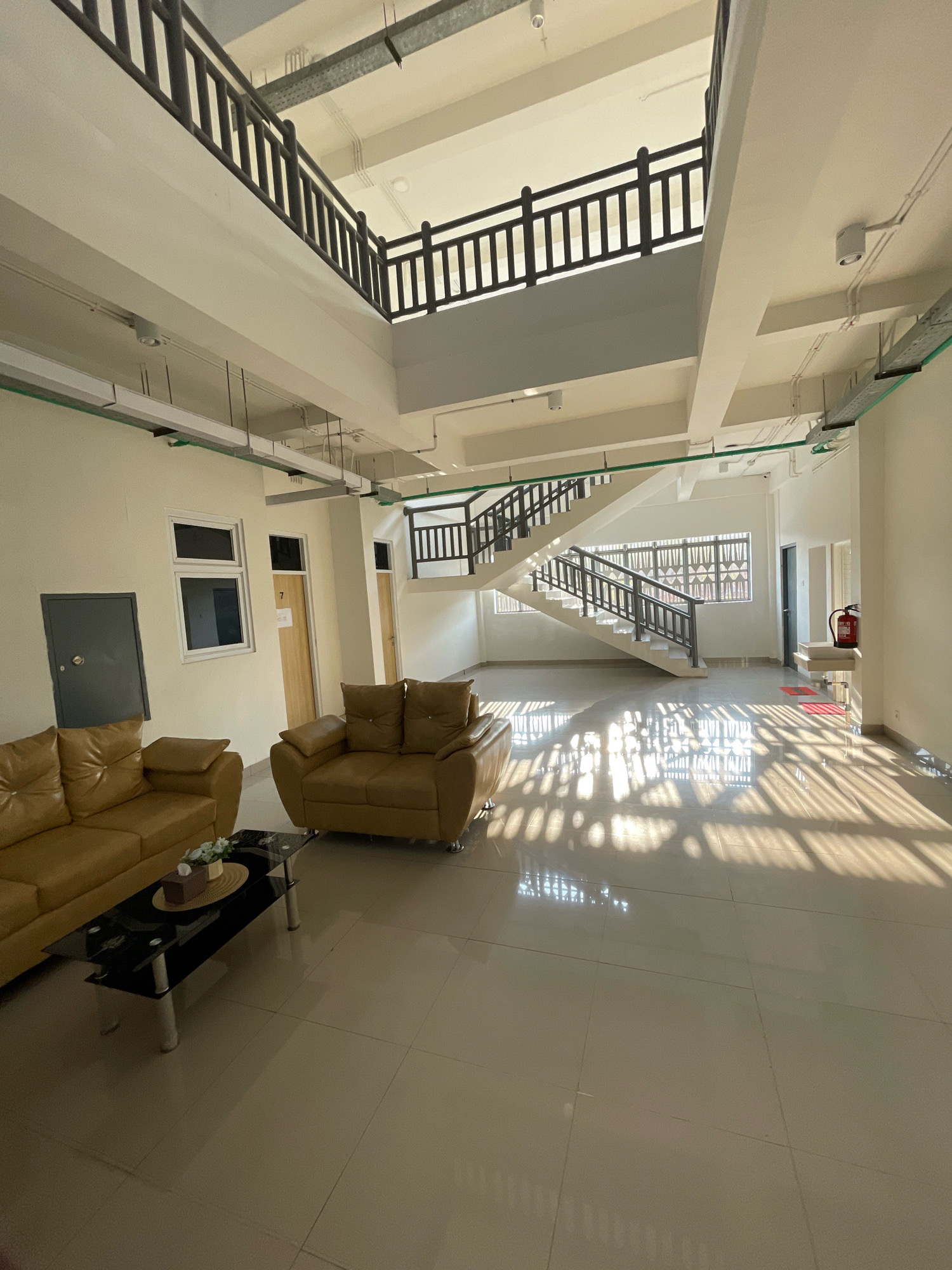 This image depicts the interior of a modern, multi-level building, likely an office or residential complex, with a pronounced architectural style featuring a prominent central staircase. The scene is bathed in sunlight, creating distinct patterns of light and shadow on the polished tiled floor, suggesting a time of day with strong natural illumination.
In the foreground, a light brown leather sofa and armchair are arranged around a rectangular glass-top coffee table. The table holds a tissue box and a decorative arrangement of flowers, suggesting a waiting or lounge area.
The background reveals a spacious open area with a white, open-riser staircase with dark railings, leading up to a second level that overlooks the main floor. The upper level also features dark railings, creating a consistent aesthetic. Throughout the space, exposed conduit piping and lighting fixtures are visible on the ceiling, indicative of a functional, contemporary design.
Several doors are present, including a dark grey metal door on the left, and two wooden doors with number "7" and another unnumbered door, to the right of the windows. One door on the right side appears to lead to a stairwell, and a red fire extinguisher is mounted on a stand near it, along with a small red mat. The walls are painted a light, neutral color.
The overall impression is one of a clean, well-lit, and functional space. There are no people visible in the image. The city of Boyangapa, Indonesia is not explicitly depicted in the image itself, but this interior scene is presented within that geographical context.