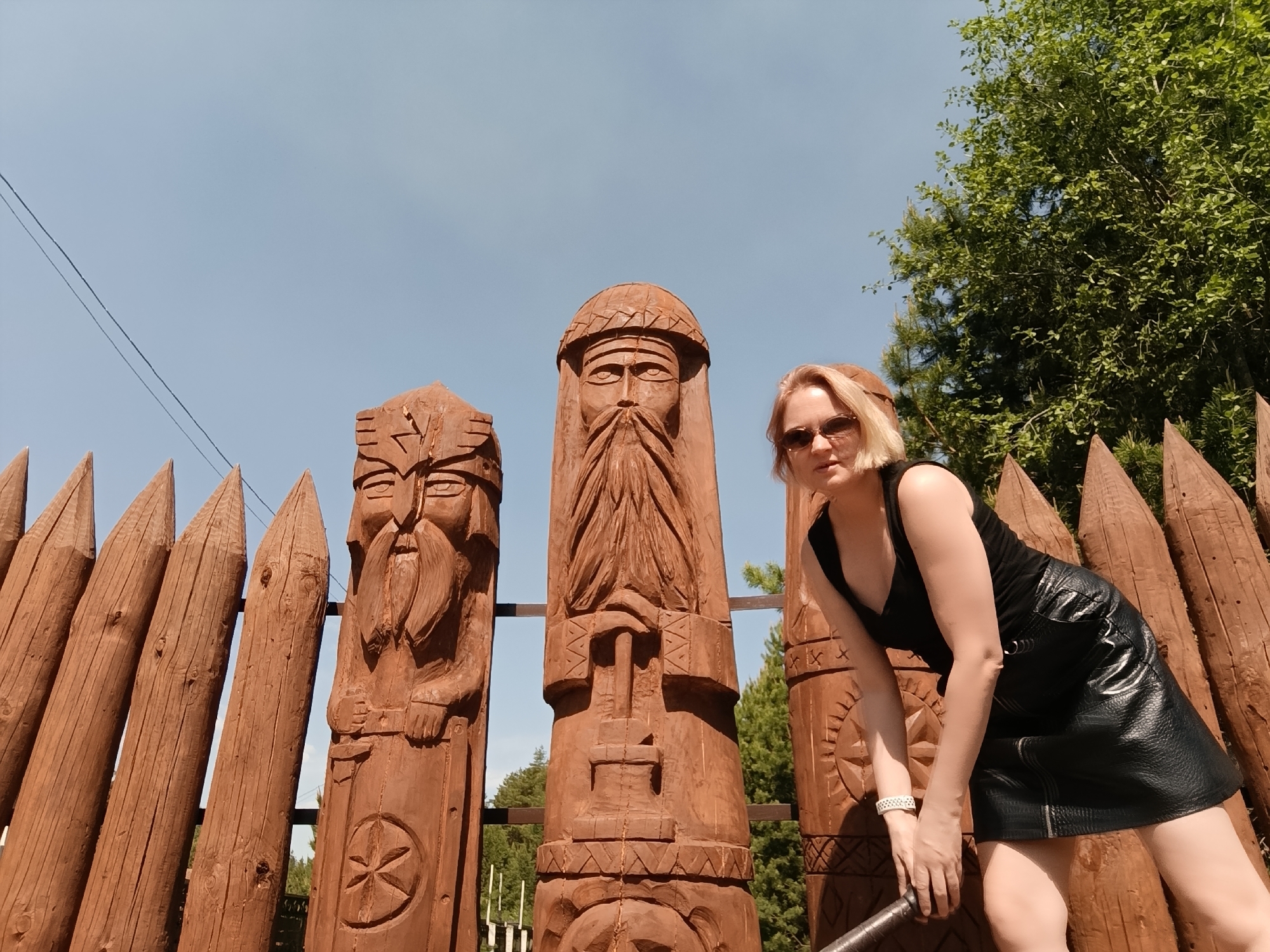 The image shows a woman in a black tank top and a black leather skirt posing with wooden carvings in Prikamskii, Russia. The carvings are of stylized human faces with beards, set against a backdrop of sharpened wooden logs, reminiscent of a palisade. To the right, a tree with green foliage is visible. The sky is clear and blue. The woman is wearing sunglasses and a bracelet, and appears to be leaning on a staff or rod. The carvings are detailed, and their brown color contrasts with the light blue sky.