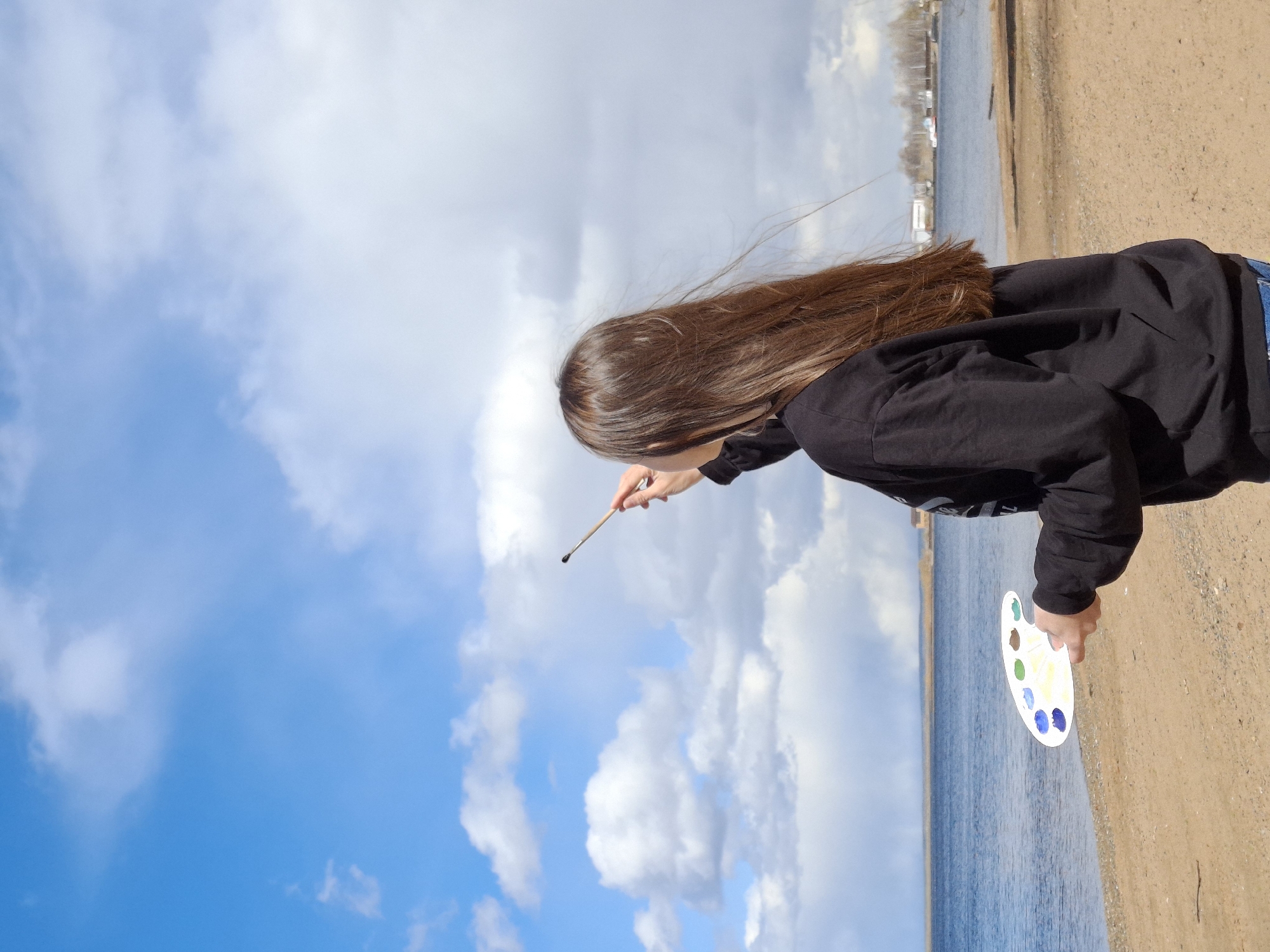 An outdoor, brightly lit daytime scene captures a person engaged in painting by a body of water. The subject, likely female, is viewed from behind, featuring long, straight brown hair. She wears a black long-sleeved top and blue jeans, partially visible.
In her right hand, she holds a paintbrush, extended outwards and upwards towards the sky, suggesting the act of painting the clouds or the expansive blue. Her left hand holds a white, oval-shaped artist's palette, which contains several distinct watercolor paints in shades of blue, green, yellow, white, and purple.
The setting consists of a sandy or dirt shoreline in the foreground, bordering a wide expanse of calm blue water, likely a lake or a broad river. Across the water in the distance, faint outlines of structures or buildings are discernible along the far bank. The sky above is a vibrant blue with scattered white, fluffy clouds, indicative of clear and pleasant weather.
The image conveys a sense of artistic creativity and a connection with nature. No discernible text is visible. The scene is set in Chaikovsky, Russia.