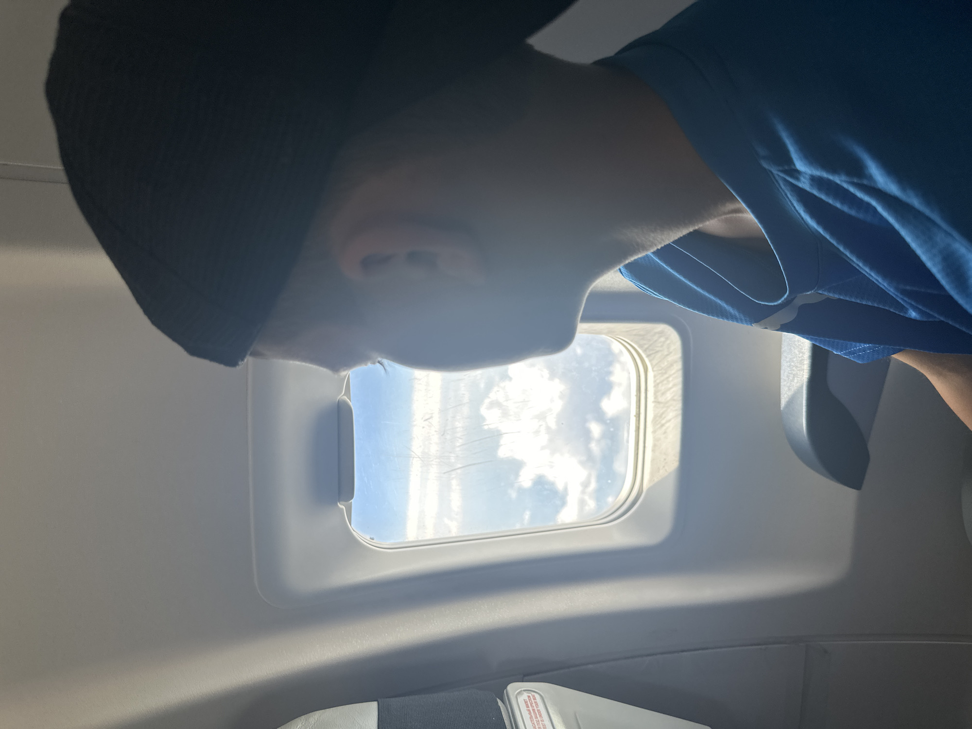 A young person wearing a black baseball cap and a blue t-shirt looks out of an airplane window. The person's face is partially obscured by the cap and the angle. The window reveals a view of white clouds against a blue sky. The interior of the airplane cabin is visible, including the window frame, a seatback, and the armrest of a seat. The lighting suggests it is daytime. There is no indication of the location being in the city of Maima, Russia.