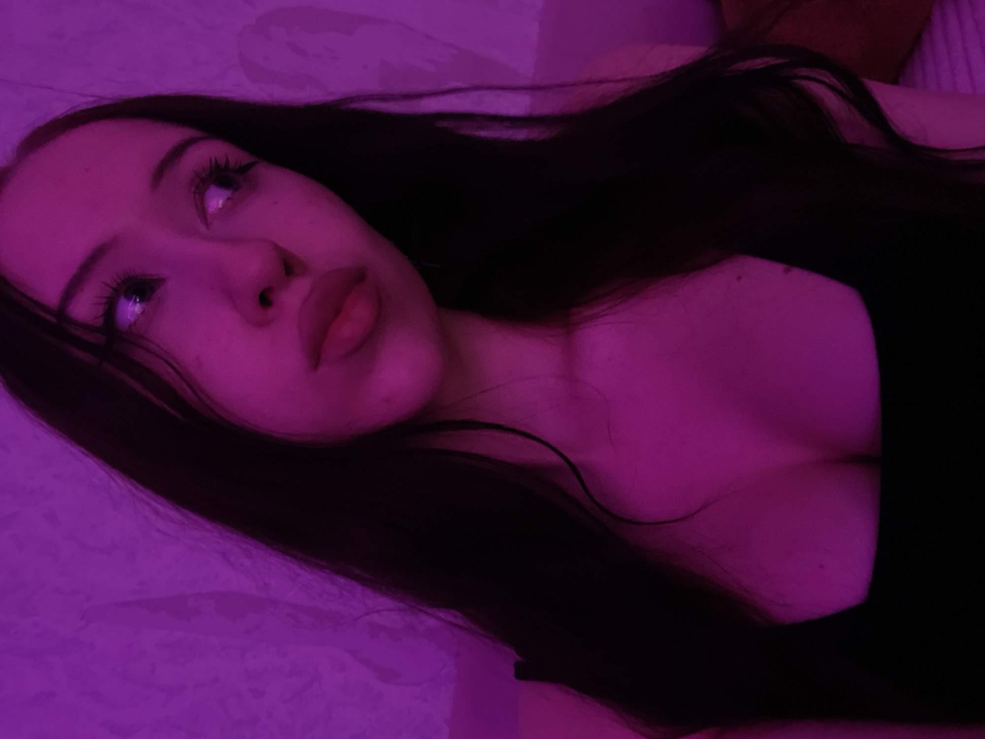 A young woman with long, dark hair is shown in a close-up portrait. She is looking to her right, with her lips slightly parted. She appears to be wearing a black top that reveals her cleavage. The lighting is a vibrant purple, casting a strong hue over the scene and highlighting the texture of the wall behind her, which appears to be a textured wallpaper. The woman's skin has some freckles visible. The image is taken indoors. There is no information about the location in Votkinsk, Russia.