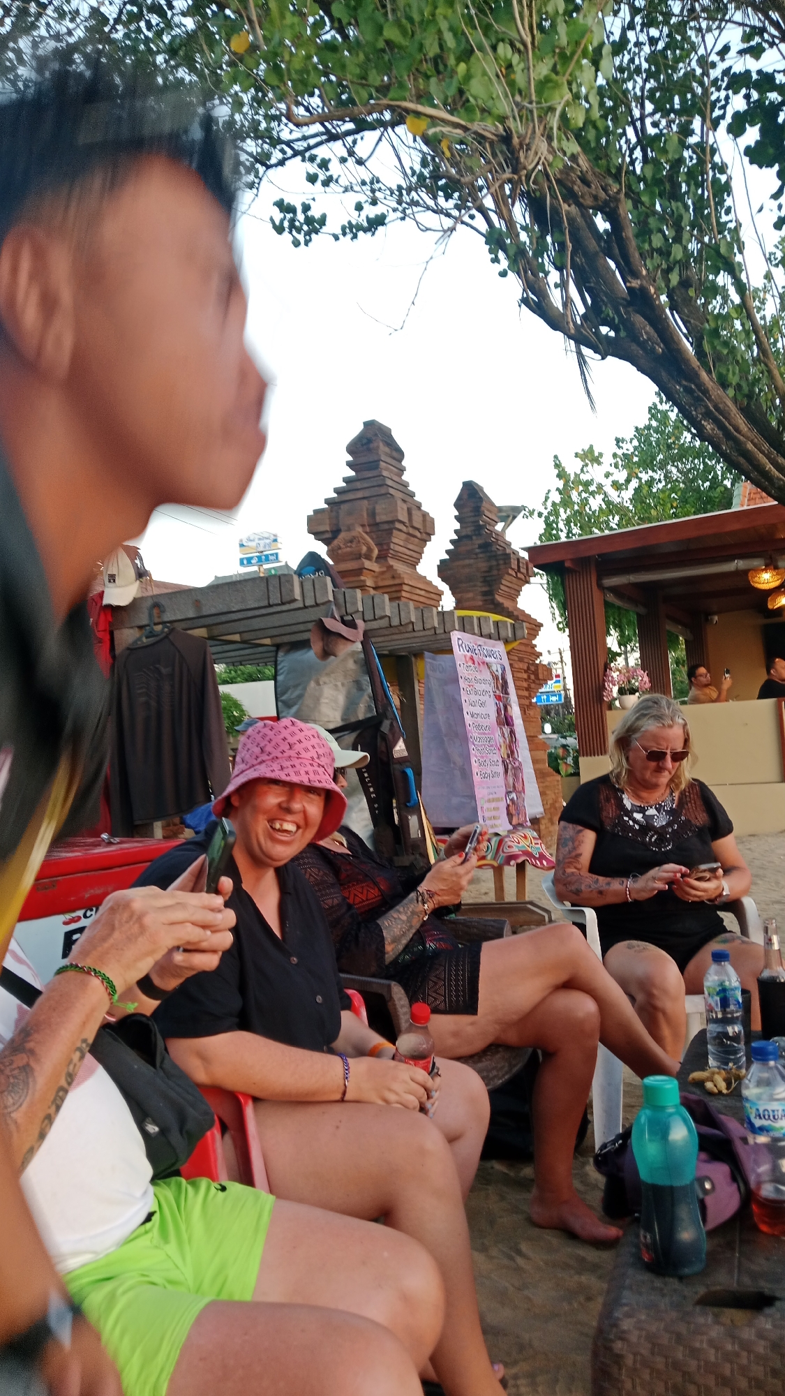 An outdoor scene in Kuta, Indonesia features a group of people seated at a table on a sandy surface, likely a beach or beachfront area, with a tree providing shade. In the foreground, a man's blurred profile is visible on the left. To his right, a woman wearing a pink bucket hat and a black top is laughing and holding a mobile phone. Beside her, another woman with blonde hair, sunglasses, and a black top is looking at her phone. The background displays a building with traditional Balinese architectural elements, featuring tiered stone structures. A sign with text, possibly a menu or service list, is also visible. Several bottles of water and a bottle of Coca-Cola are on the table. Clothing items are displayed on a rack behind the people. The overall atmosphere suggests a casual gathering in a tropical setting.