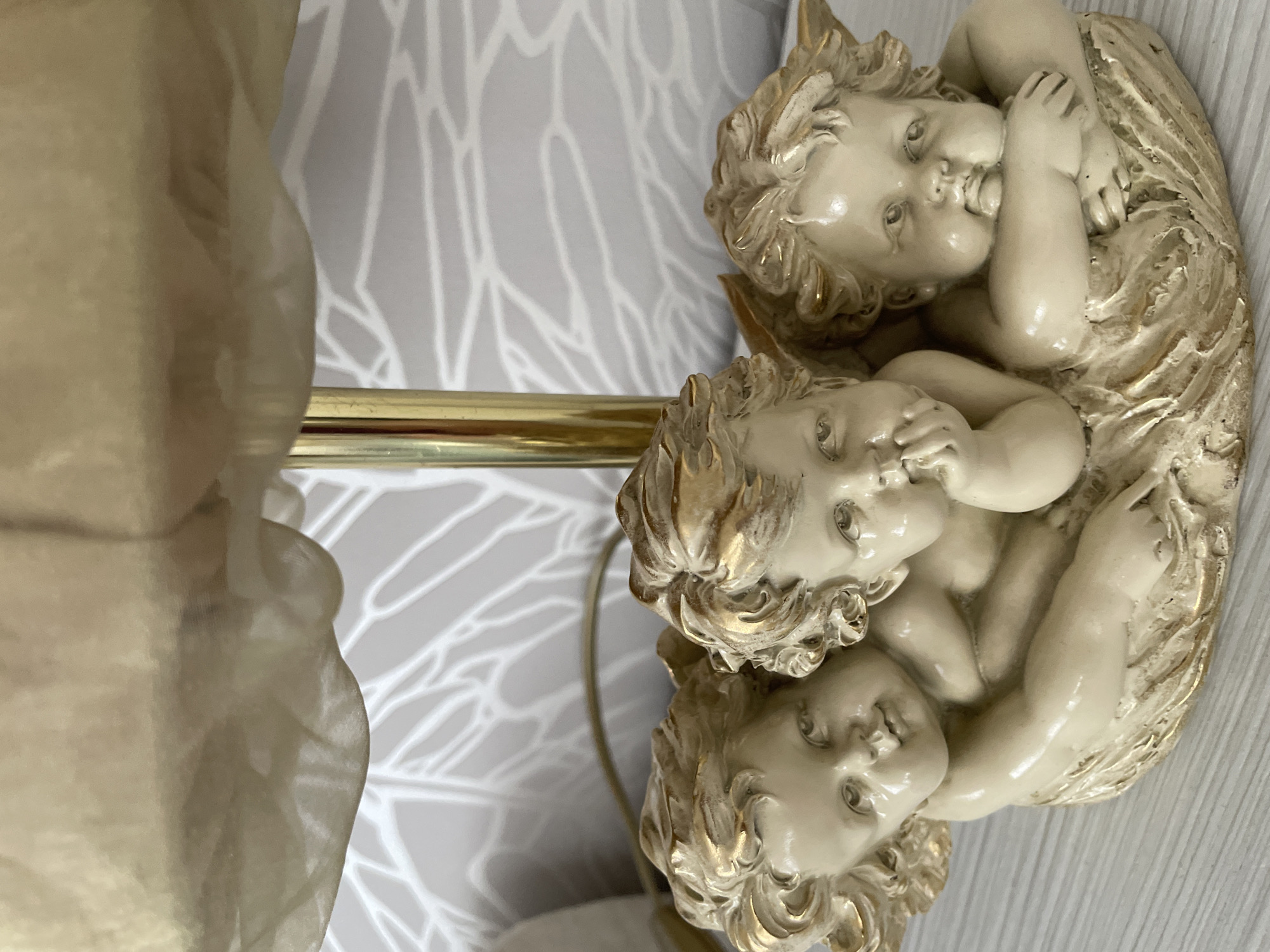 The image depicts a decorative lamp featuring three cherub figurines nestled together at the base. A gold-colored metal rod rises from the cherubs and is connected to a translucent, off-white shade, likely made of a textured material. The background shows a light grey wallpaper with a white linear pattern. The objects appear to be indoors and based on the lighting conditions likely artificially illuminated. There is insufficient information to determine a specific location in Лесной Пейзаж Кп (шарапово, Russia.