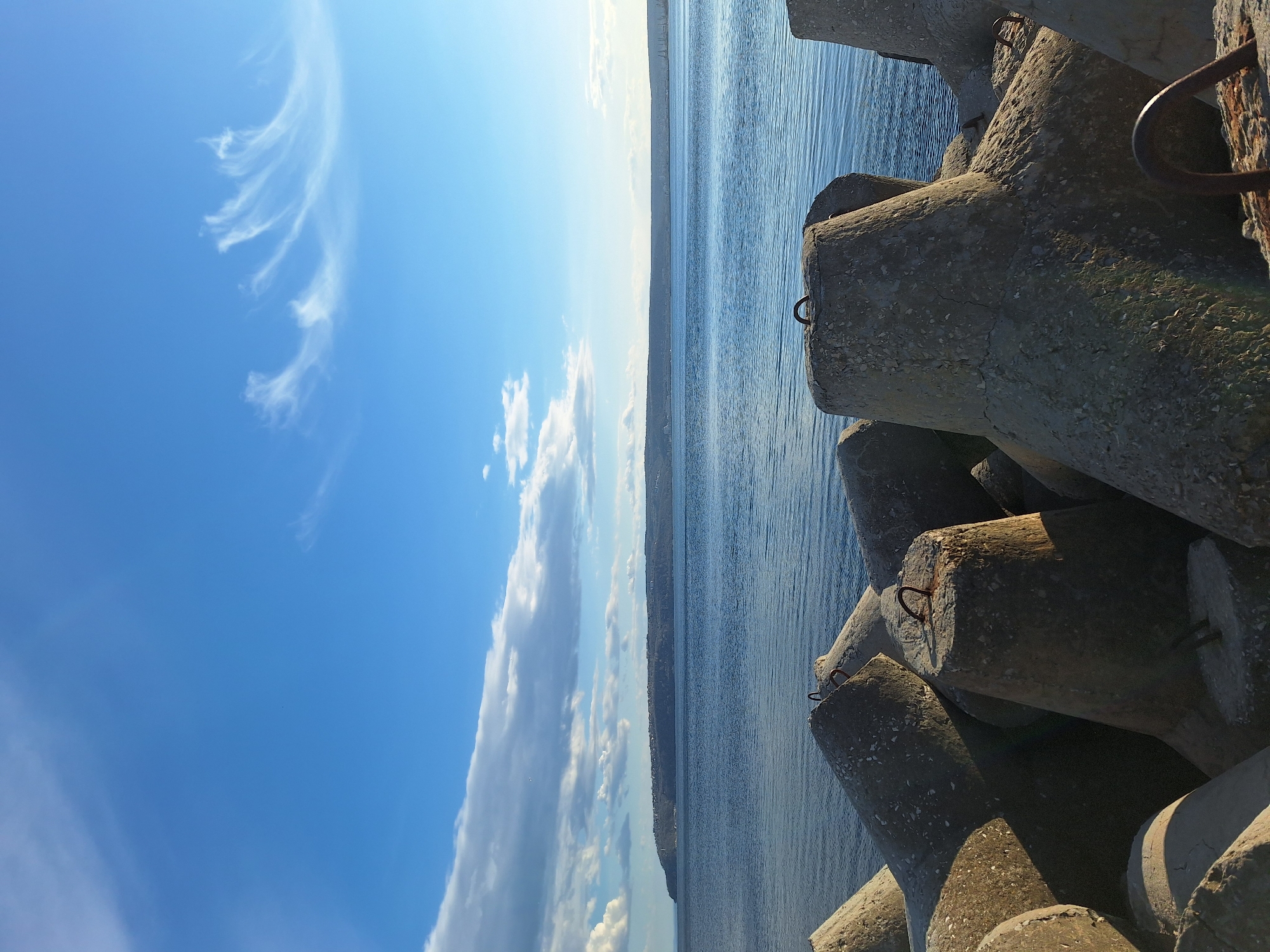The image shows a scene at a Varna, Bulgaria breakwater. Tetrapod concrete structures are stacked on the right side of the frame. The sea extends to the middle of the image, with a coastline visible in the distance. On the left, the sky is blue with scattered clouds.