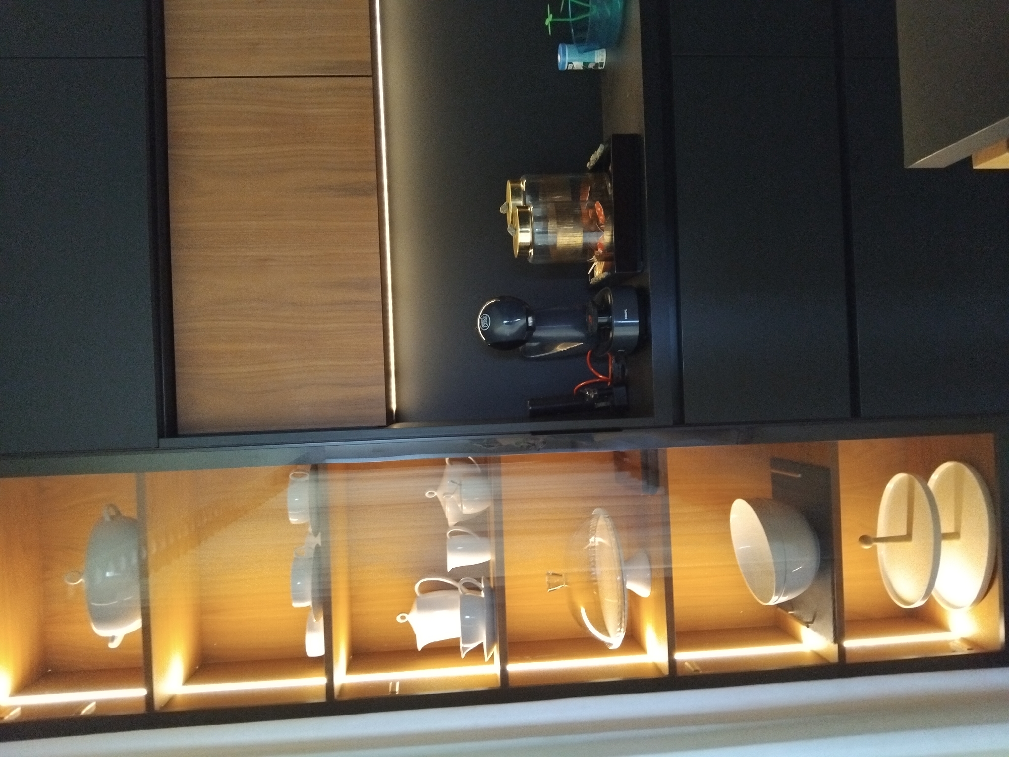 In an indoor scene, a well-lit display cabinet with wooden shelves is filled with white ceramic tableware, including bowls, cups, and teapots. The shelves are illuminated by vertical LED lights. To the right of the cabinet is a dark countertop featuring a black coffee maker, two glass jars with gold lids containing what appear to be capsules or coffee pods, and a small blue bottle. Above the countertop are dark grey cabinets and a section of wooden cabinetry. The bottom right of the image shows the corner of a dark table or countertop. The overall environment suggests a modern kitchen or dining area. There are no people visible in the image.