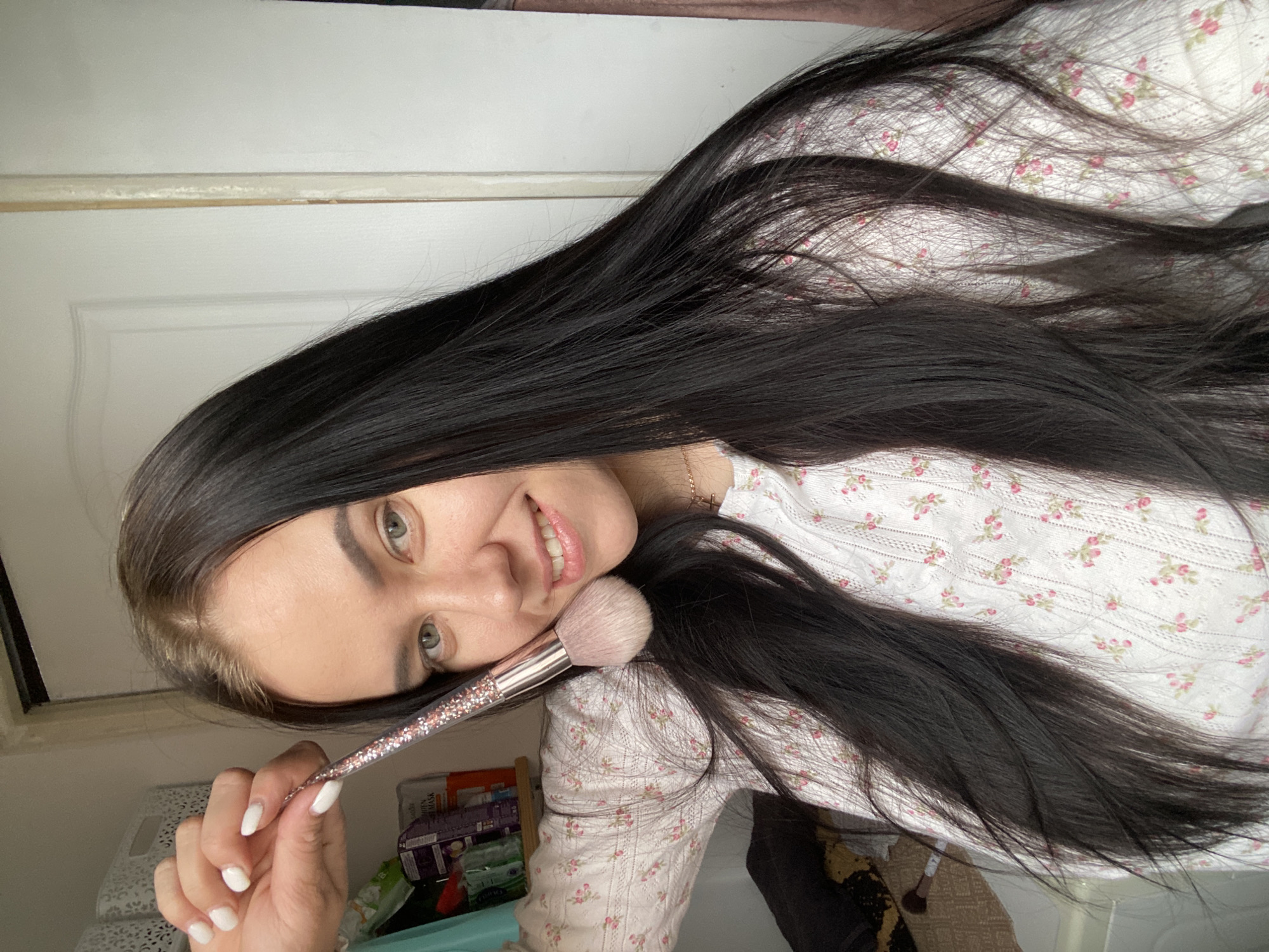 A woman with long, dark hair is holding a large makeup brush in her right hand, positioning the brush's head against her cheek. She is smiling and looking at the camera. She is wearing a light-colored floral-patterned top. The background includes a white door and miscellaneous items on a shelf or surface, some of which appear to be personal care products. The image's location is Chaikovsky, Russia.