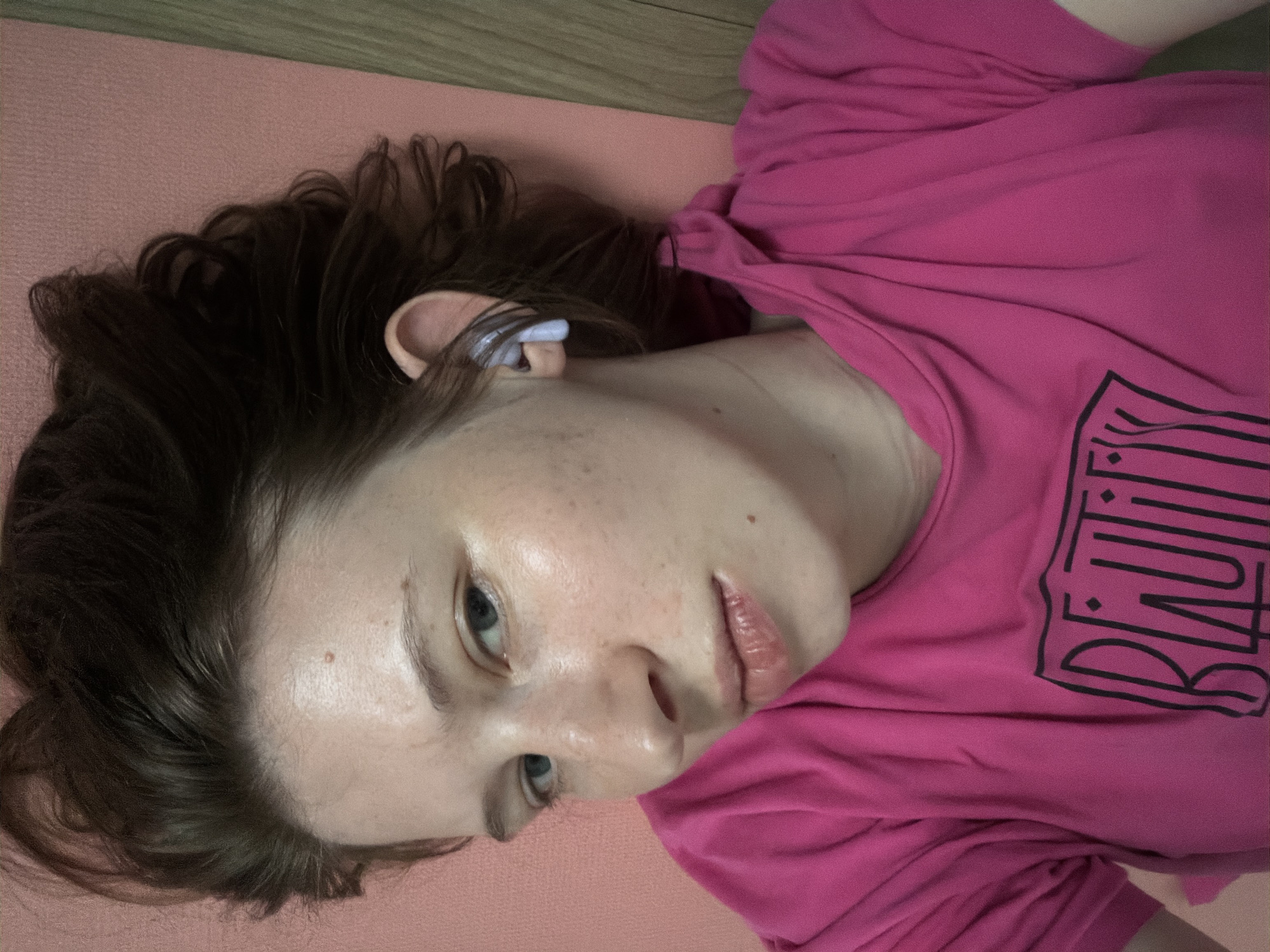 A young woman with brown hair, wearing a bright pink hoodie with "BEAUTY" written in black on it, is lying on a pink surface, possibly a yoga mat. She has blue eyes and is wearing a white wireless earbud in her right ear. The background shows a wooden surface to the right. The image appears to be a selfie taken from a close-up, overhead perspective. The lighting suggests an indoor setting. There are no discernible landmarks or indicators to pinpoint the location to Dongducheon-si, South Korea.