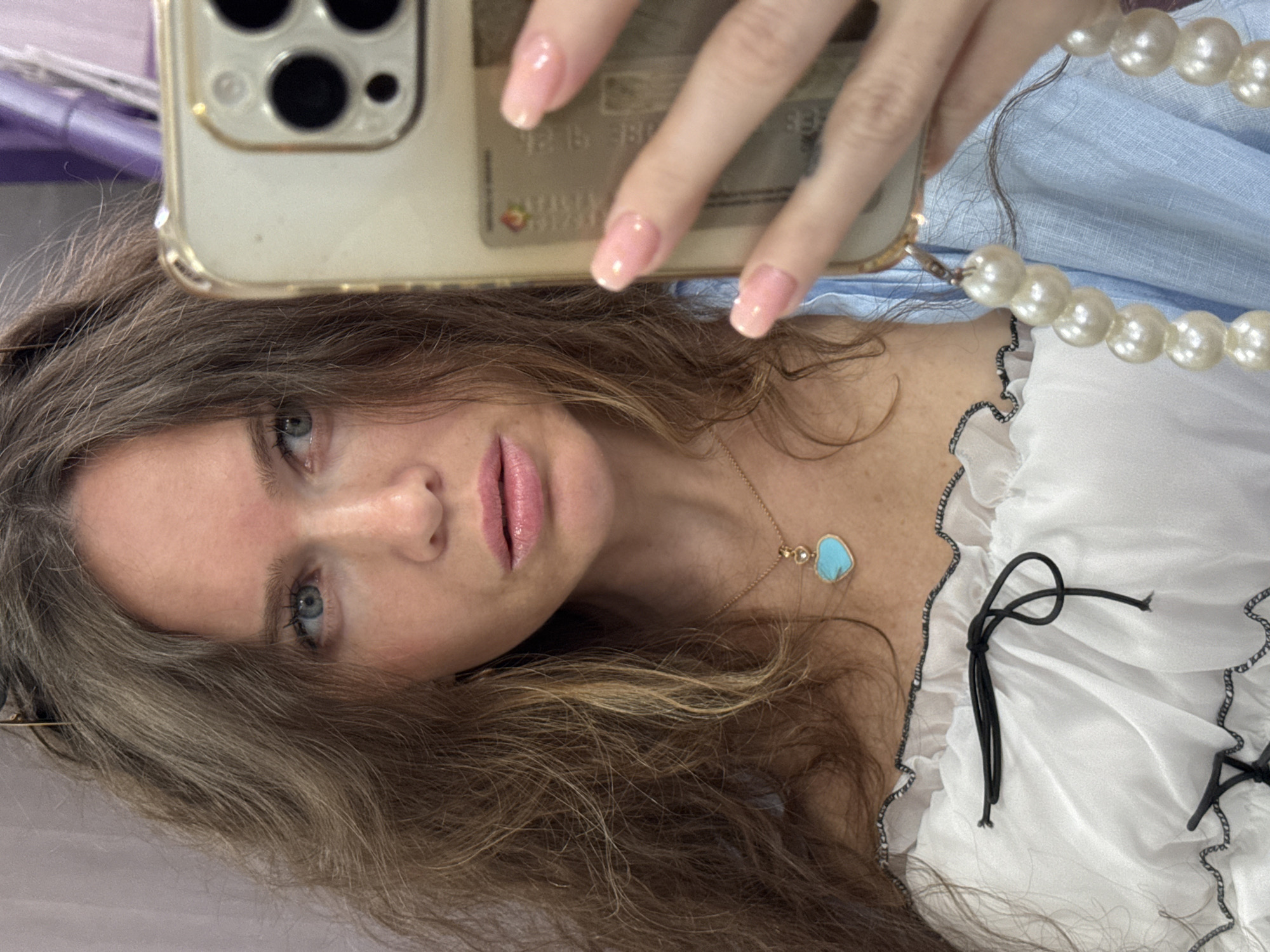 This is a vertical, close-up, selfie-style photograph of a young woman. She is holding a smartphone with multiple camera lenses in her right hand, which is positioned in the upper portion of the frame. Her left hand is also visible, with light pink, glossy fingernails. The woman has long, wavy brown hair that frames her face. She is wearing a white, ruffled blouse with black trim and black tie closures. A gold necklace with a light blue, heart-shaped pendant is visible on her chest. The background is blurred, but appears to be an indoor setting, possibly a room with purple and white elements. The lighting suggests it is daytime. The woman's expression is neutral, and the overall mood is casual and personal. There is no visible text in the image, although a credit card is partially visible through the back of the phone case. The image was taken in Moscow, Russia.