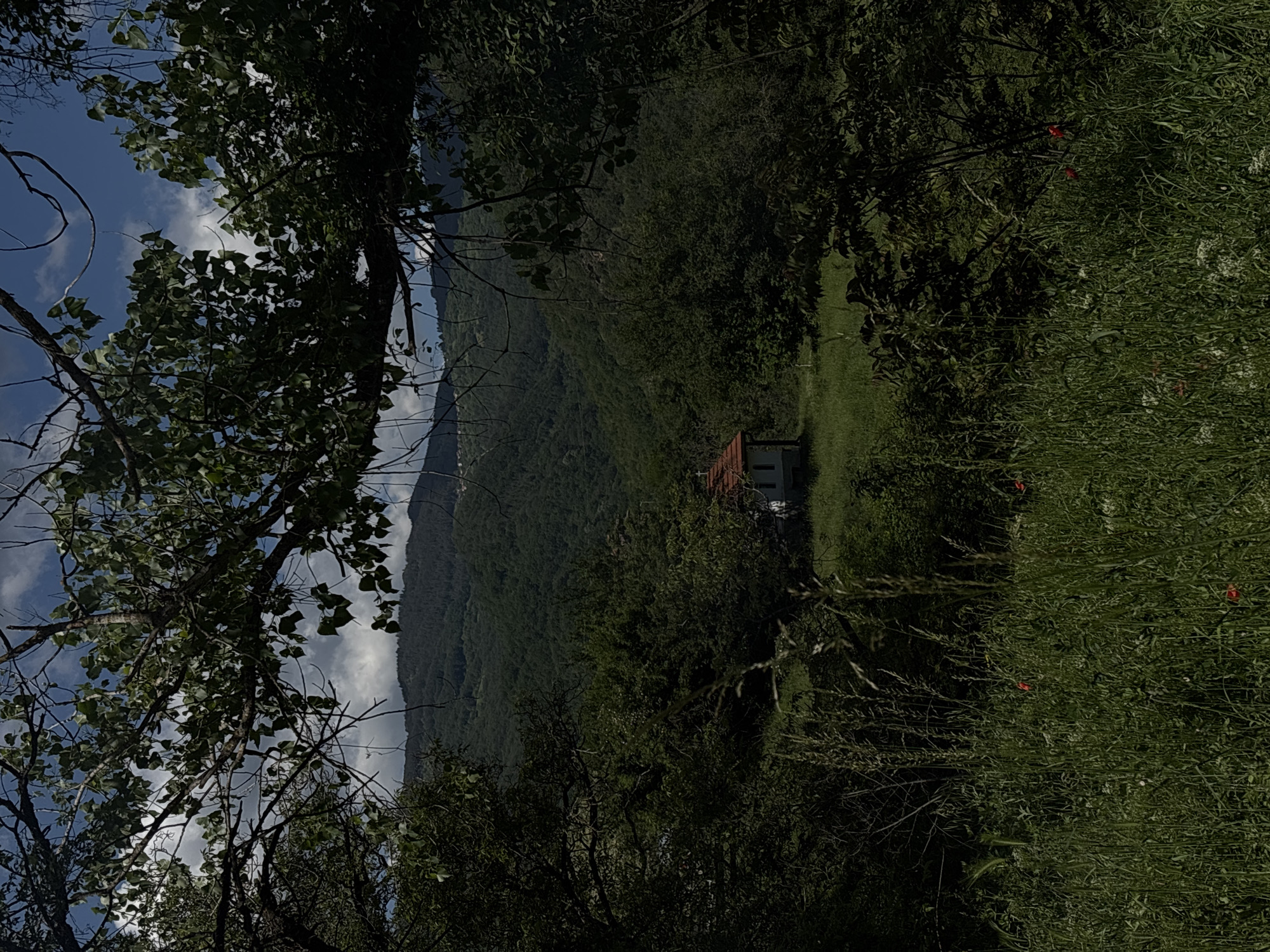 The image depicts a landscape with a small building nestled in a green meadow, framed by trees and foliage. In the background, there is a mountain range under a sky with scattered clouds. The foreground has an abundance of vegetation, suggesting a natural, possibly rural, environment. The scene seems to be a viewpoint looking down on the building and the surrounding landscape. The location is stated to be in the city of Unknown, Unknown.