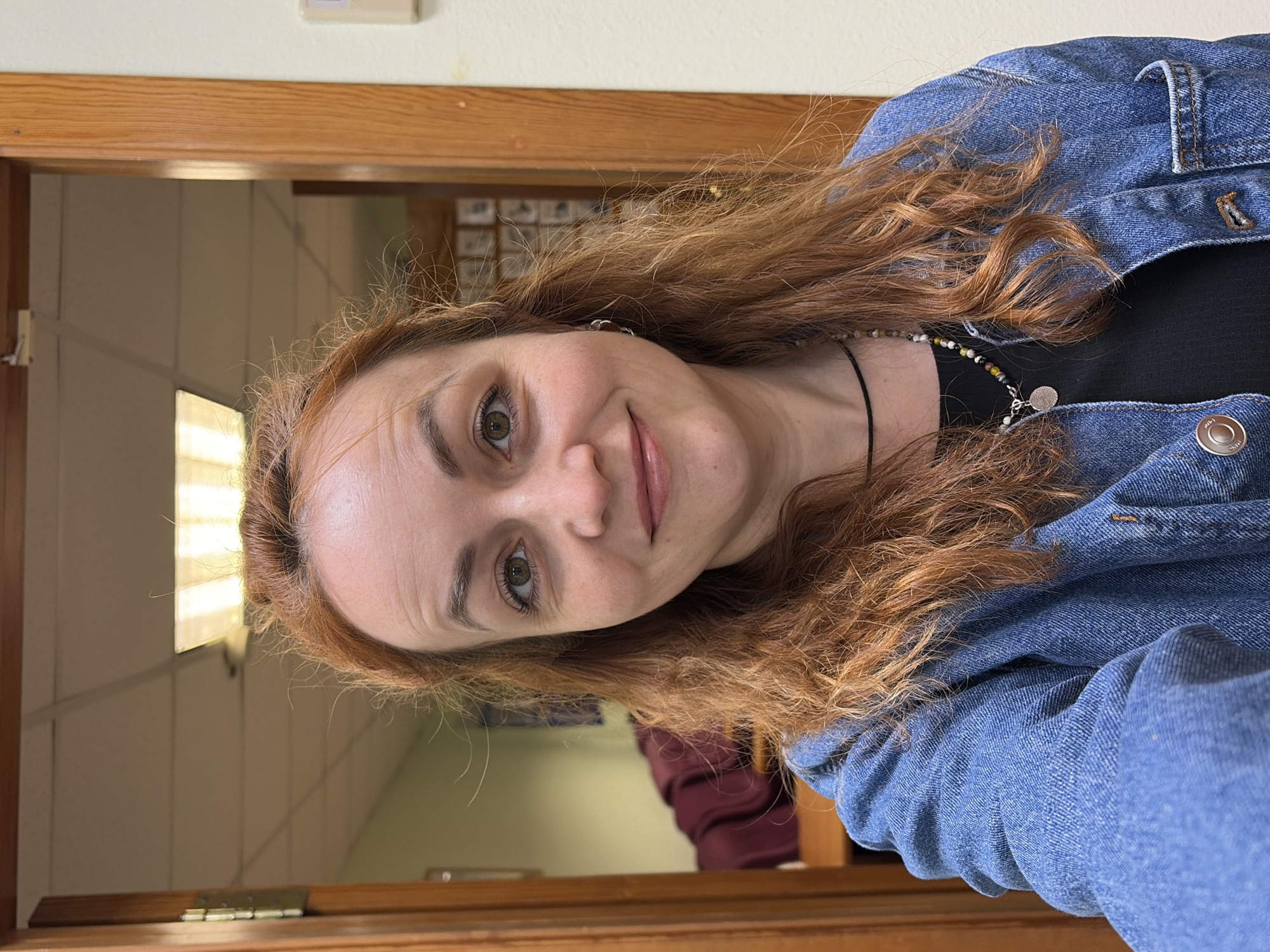 A woman with auburn curly hair, light skin, and green eyes is centered in the frame, wearing a denim jacket over a black top and a necklace with beads and a silver pendant. She is facing the camera with a slight smile. The background reveals a glimpse of an interior space through a wooden-framed doorway. A fluorescent light fixture is visible, suggesting an office environment, with a cream-colored wall above the doorframe. Documents or images are affixed to a wall in the interior space. The image's location cannot be determined from this image.