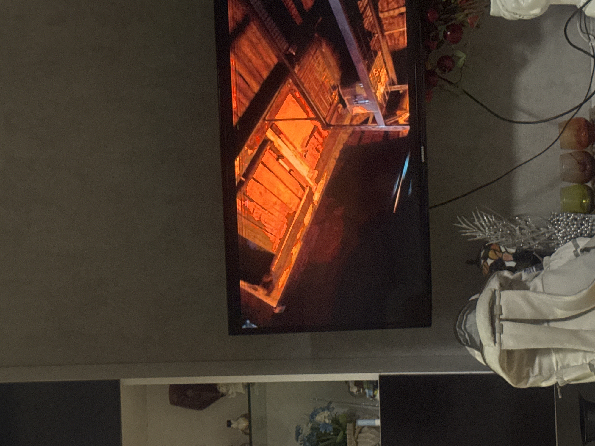 A television screen displays an interior view of a building, appearing to be wood-structured with orange-toned lighting, possibly a historical building or a traditional residence. The television is situated on a stand, and out of focus, there are decorations and household items nearby. The environment is indoors, with neutral-colored walls. There are no people visible in the frame. It's difficult to confirm the exact location as Tbilisi, Georgia without further contextual clues.