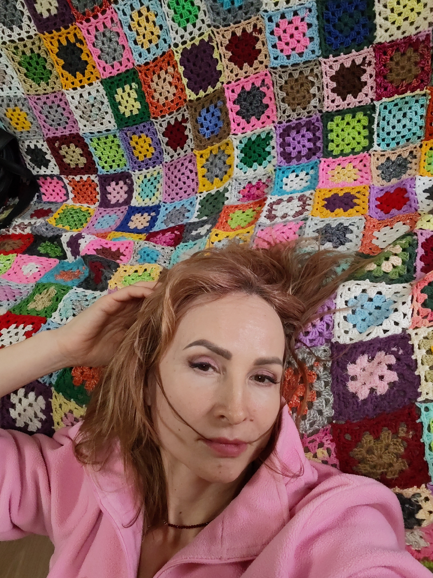A woman with auburn hair, wearing a pink fleece jacket and a necklace, is lying on a multicolored crocheted blanket composed of many small squares. Her right hand is raised towards her hair. The image was likely taken indoors. There is no contextual information to determine the location in the city of Chaikovsky, Russia.