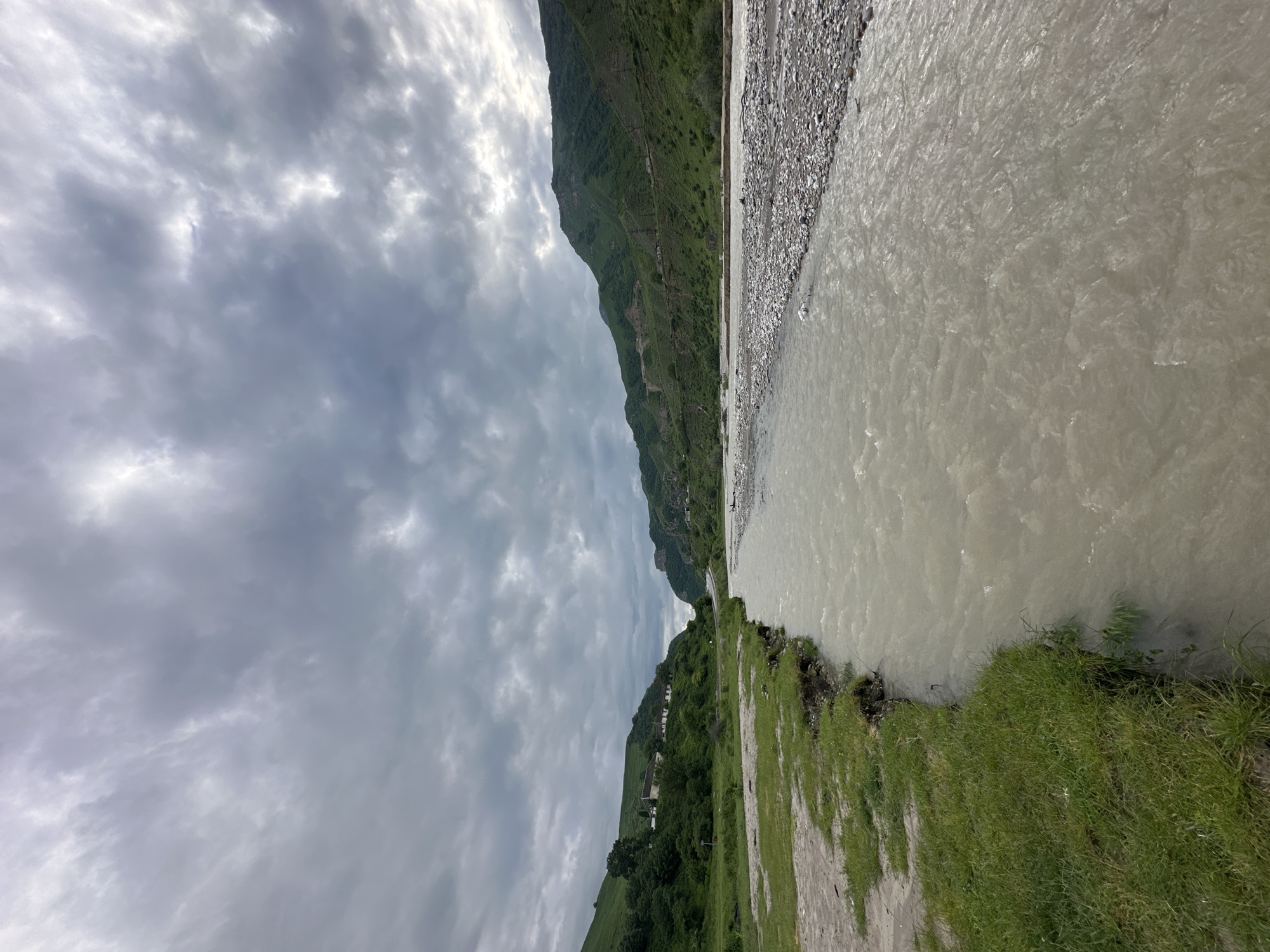 The image depicts a wide, muddy river flowing through a valley. The riverbed on the right side of the image is covered with pebbles. Lush green hills rise on both sides of the valley, with scattered houses and trees visible on the slopes. The sky is overcast with dramatic, grey clouds. The perspective is from ground level, looking downstream. The scene appears to be in a rural or mountainous area. Based on the provided context, this is the river in Bedyk, Russia.