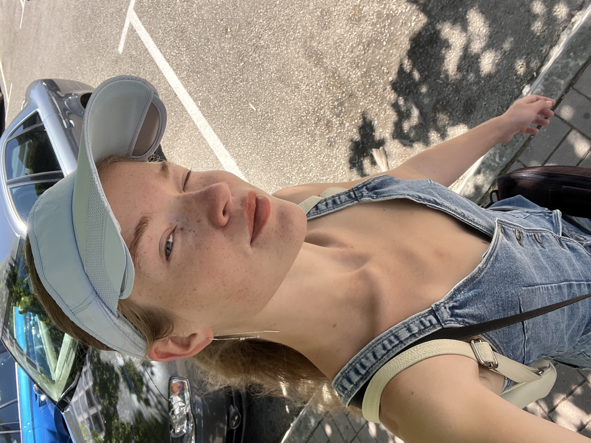 A young woman with freckles and blonde hair pulled back is taking a selfie outdoors. She is wearing a light blue visor, a denim overall dress, and a white purse with a strap. She is also wearing a long, delicate earring in her left ear. Behind her, a grey car is parked in a lot with white parking lines. The ground is asphalt with some shadows from trees cast upon it. The scene appears to be a sunny day in a city.