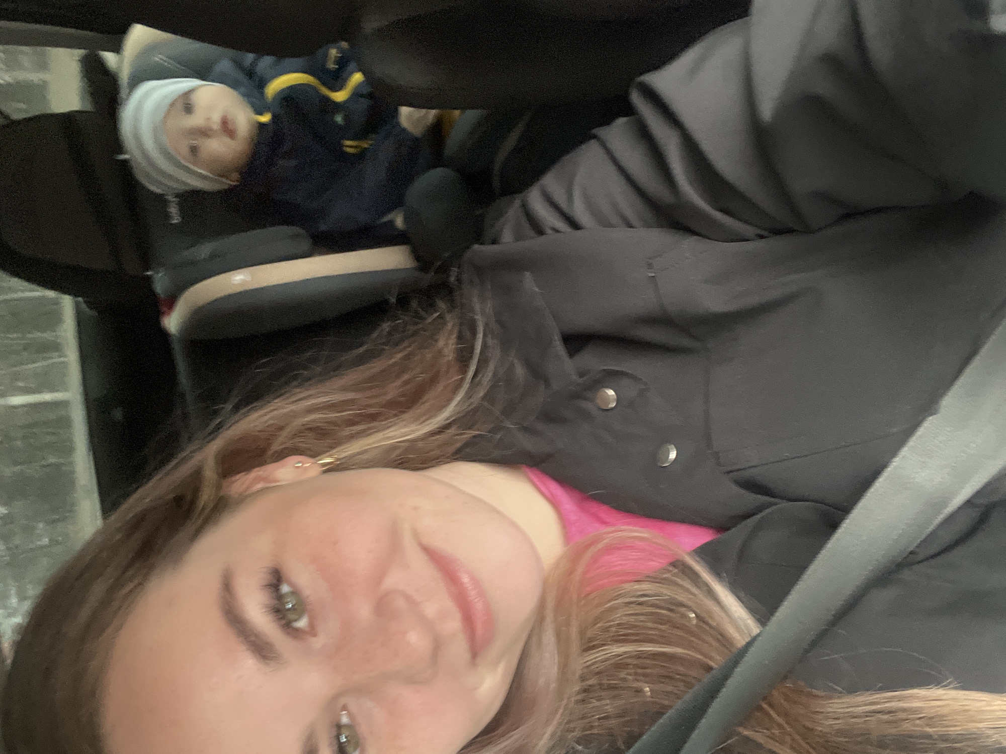 A selfie taken in a car. A woman is wearing a black jacket over a pink shirt. She has light brown hair and green eyes. A child wearing a blue and yellow jacket and a white and grey hat is visible in a car seat behind her. The interior of the car is black and grey. No information suggests the location is Chaikovsky, Russia.