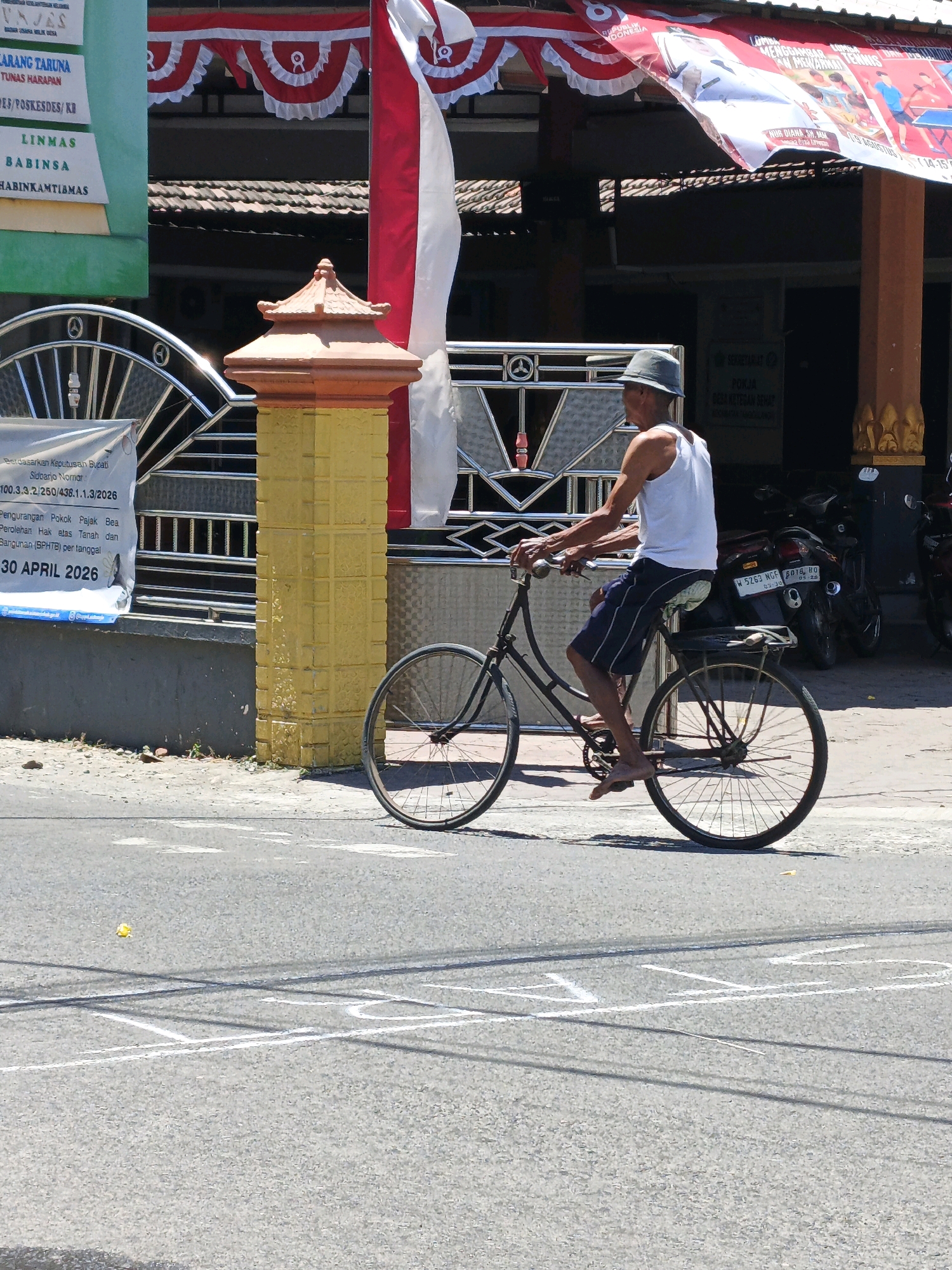A middle-aged man wearing a white tank top and dark striped shorts rides a black bicycle. He is wearing a light gray hat and is barefoot. The man is cycling on a paved road in the city of Ketegan, Indonesia. The road has white markings on it. Behind him, there are buildings with red and white banners hanging from them. There are also motorcycles parked near the buildings. The man is looking to his right and appears to be pedaling. The sun is shining, casting shadows on the road.