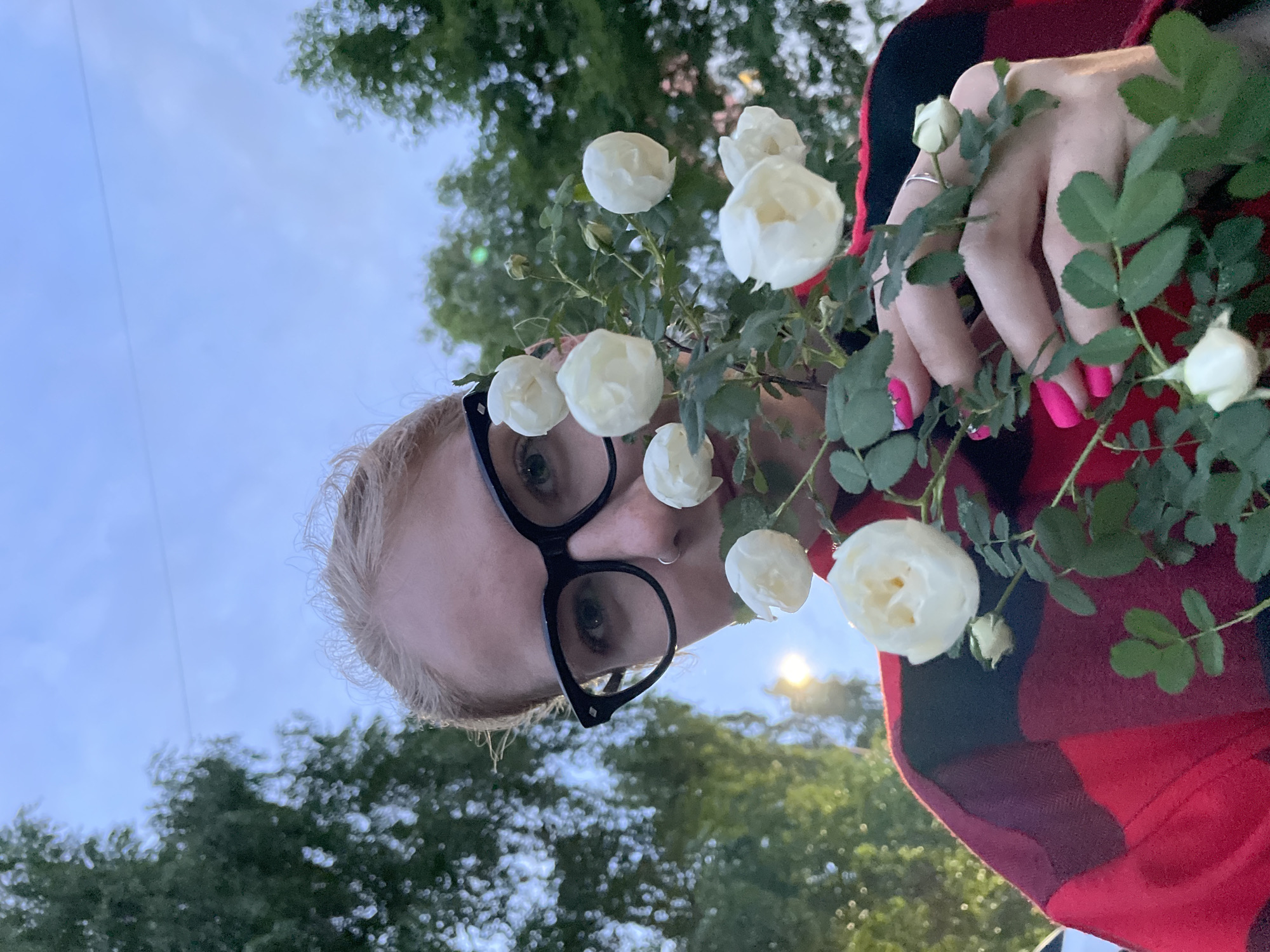 A low-angle shot depicts a person holding white roses. The individual has blonde hair, wears black-rimmed glasses, and is clad in a red and black plaid garment. Their fingernails are painted a vibrant pink. The background features trees and a blue sky. The location is unspecified, and there is no indication that the photo was taken in Izhevsk, Russia.