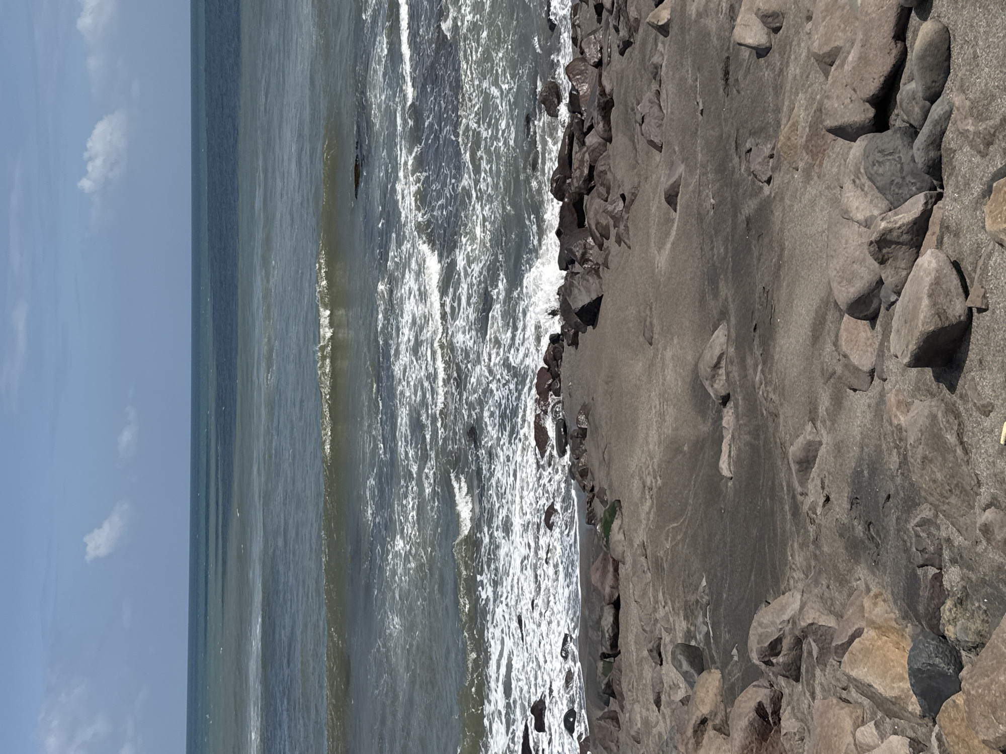 The image shows a coastal scene in Mengwi, Indonesia. The ocean is visible on the left side, with waves crashing towards the shore. The shoreline consists of dark gray sand and is lined with rocks. The sky above is partially cloudy. The image is taken at a relatively high angle, looking down at the beach. There are no people visible in the image.
