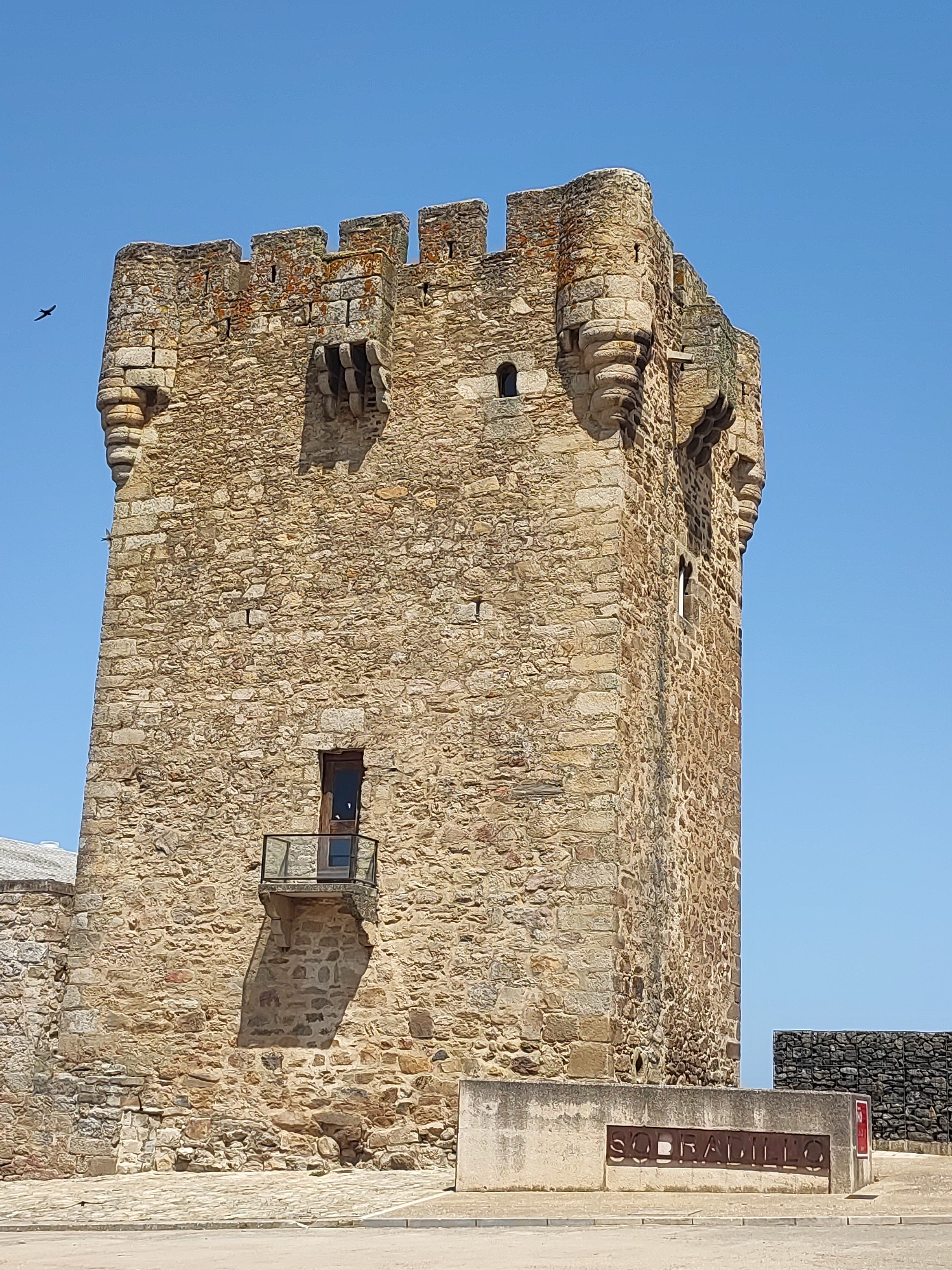 The image captures the Torre de Sobradillo, a historic stone tower located in the city of Sobradillo, Spain. The tower, constructed from rough-hewn stones of varying sizes and shades of brown and gray, stands prominently against a clear blue sky. Its crenellated parapet features square merlons and embrasures, with some sections showing patches of orange lichen. Decorative stone corbels project from the upper corners of the tower, adding architectural detail. A small, dark-paned wooden door is visible on one face of the tower, above which juts out a small balcony with a glass railing. A shadow is cast on the tower wall from this balcony. To the left of the tower, a portion of another stone structure with a light-colored roof is partially visible. In the foreground, a low concrete wall bears the name "SOBRADILLO" in cutout metal letters. Beyond the wall, a dark, gabion-style stone wall is visible. A single bird is in flight in the upper left portion of the sky. The ground in the foreground appears to be paved with cobblestones and asphalt.