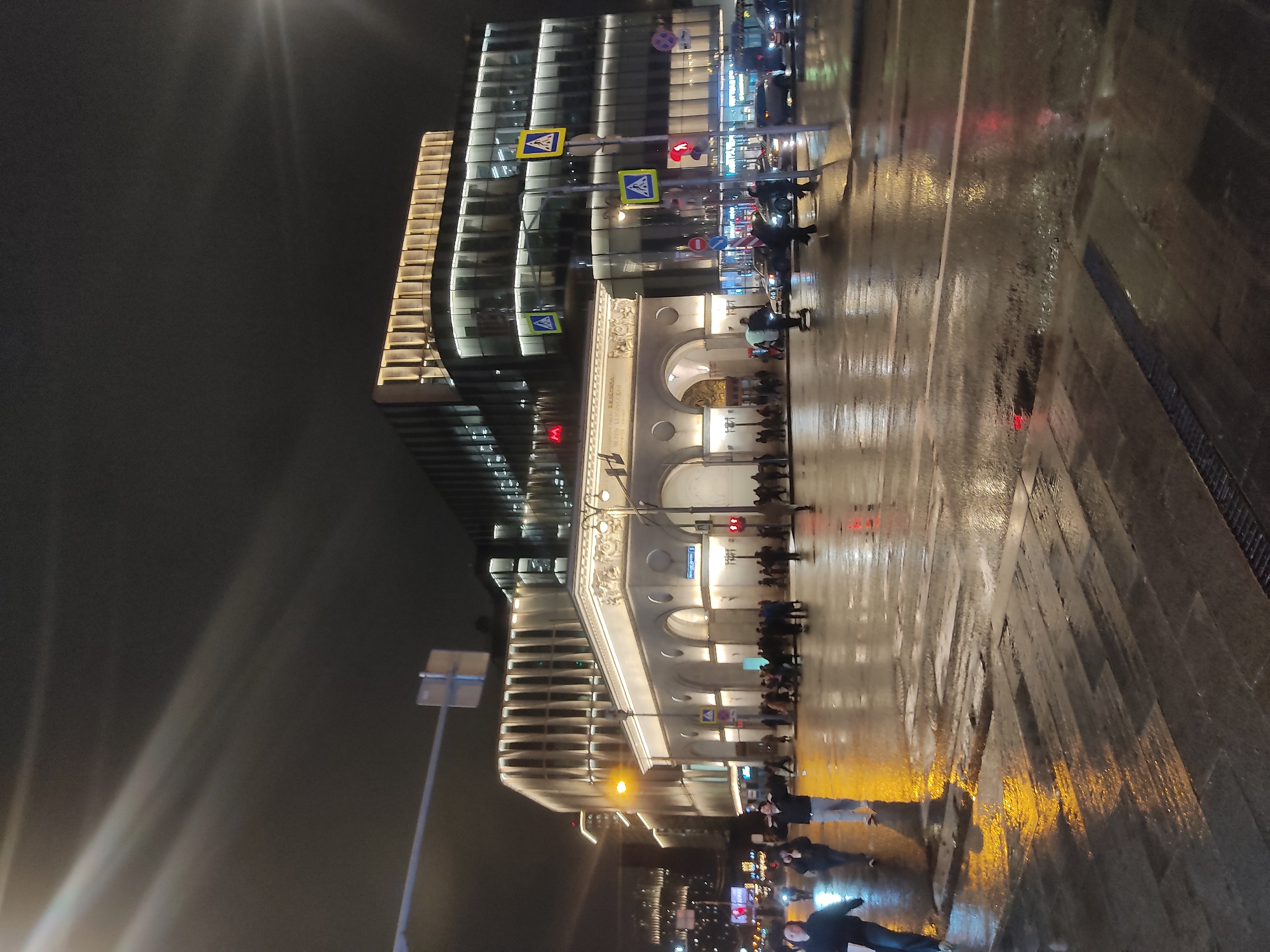 At night, a city street scene shows a modern building with glass and horizontal lighting, adjacent to an older-style, arched building with white stone. Pedestrians line the sidewalk in front of the arched building. Streetlights reflect off a wet, cobblestone crosswalk. Several road signs are visible.