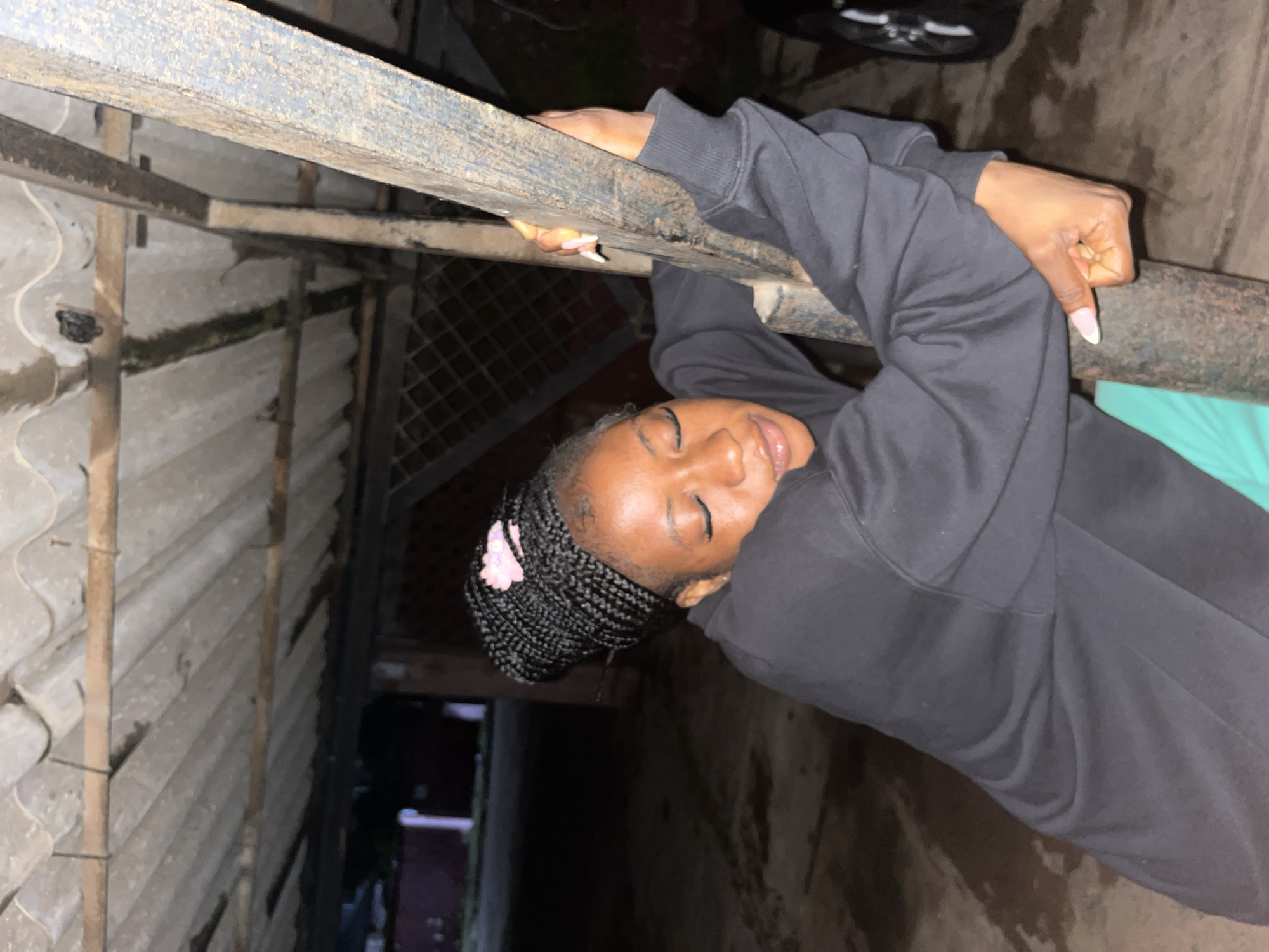 A young Black woman with her eyes closed and head tilted down is leaning against a metal pole in what appears to be a covered outdoor area. She has dark braided hair styled in cornrows, with a small pink hair clip on top. She is wearing a black hooded sweatshirt and bright green shorts. Her right arm is wrapped around the pole, and her left hand is also gripping it. The background suggests an urban environment, possibly a parking garage or an open-air structure, with a corrugated metal roof overhead and a chain-link fence visible in the distance. The ground is paved and wet, indicating recent rain. The overall lighting is dim, suggesting it is either nighttime or an indoor shaded area. Based on visual cues, the location is unlikely to be definitively identifiable as Jos, Nigeria without further information.